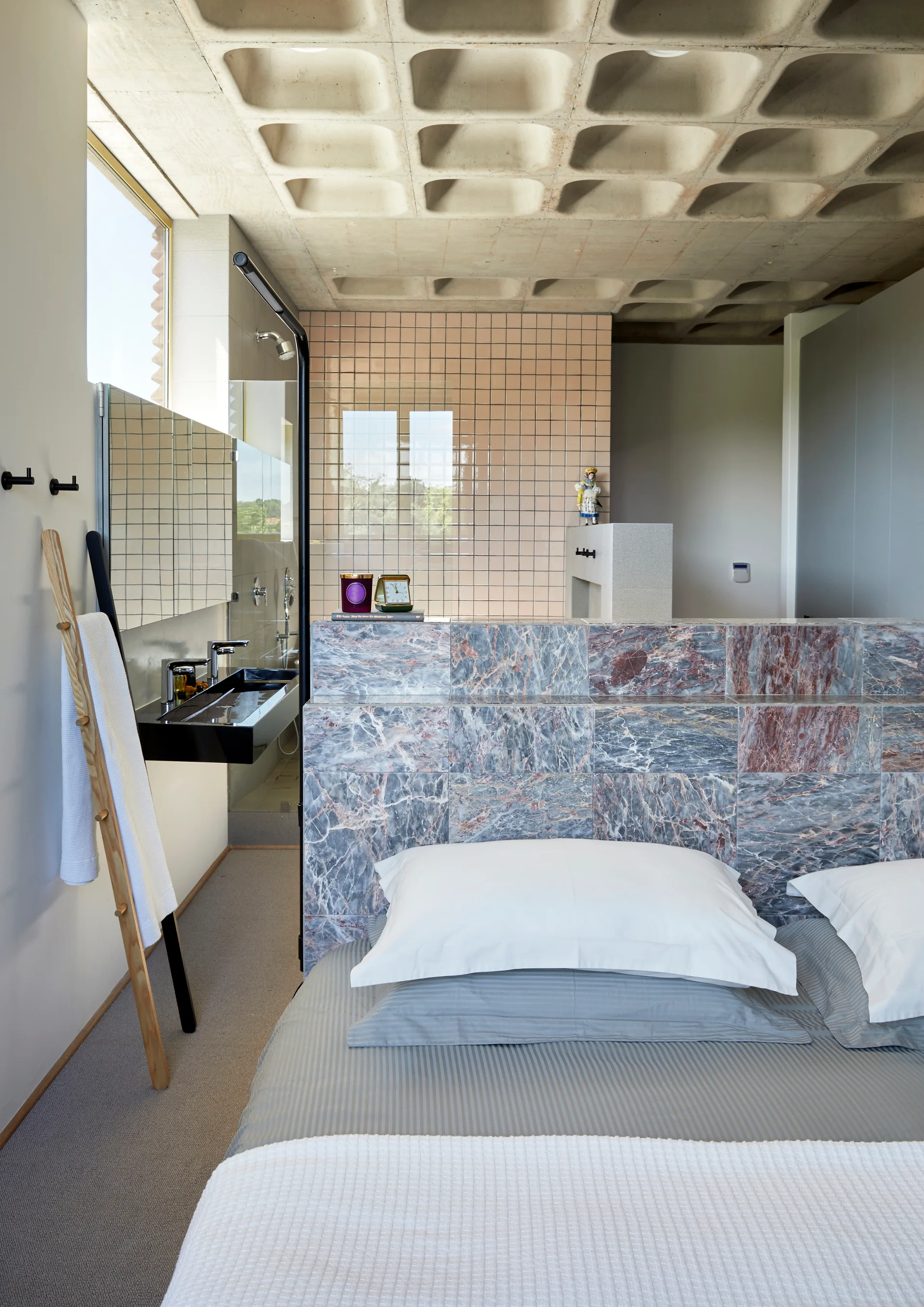 A bed with grey linens sits before a blue marble partition wall beneath an exposed concrete waffle ceiling in an open bedroom and bathroom suite.