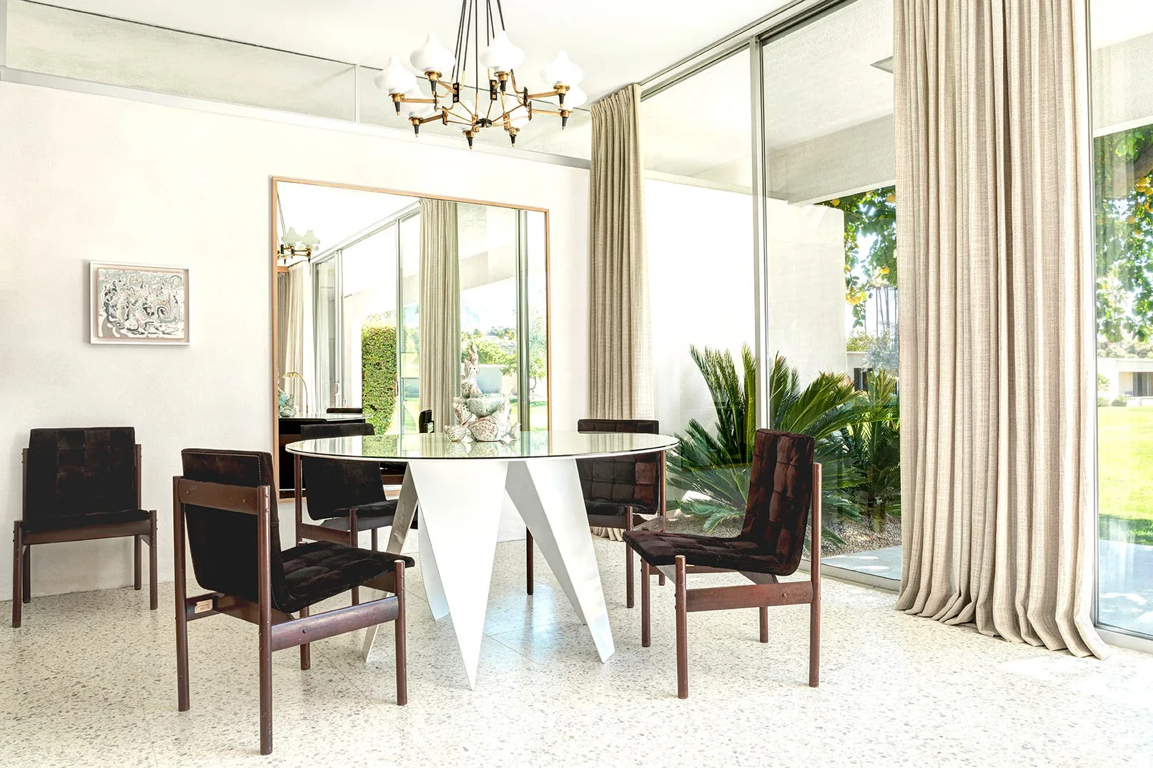 A mid-century modern dining room features a round glass table with a white geometric base, dark wood chairs, terrazzo flooring, and glass walls.
