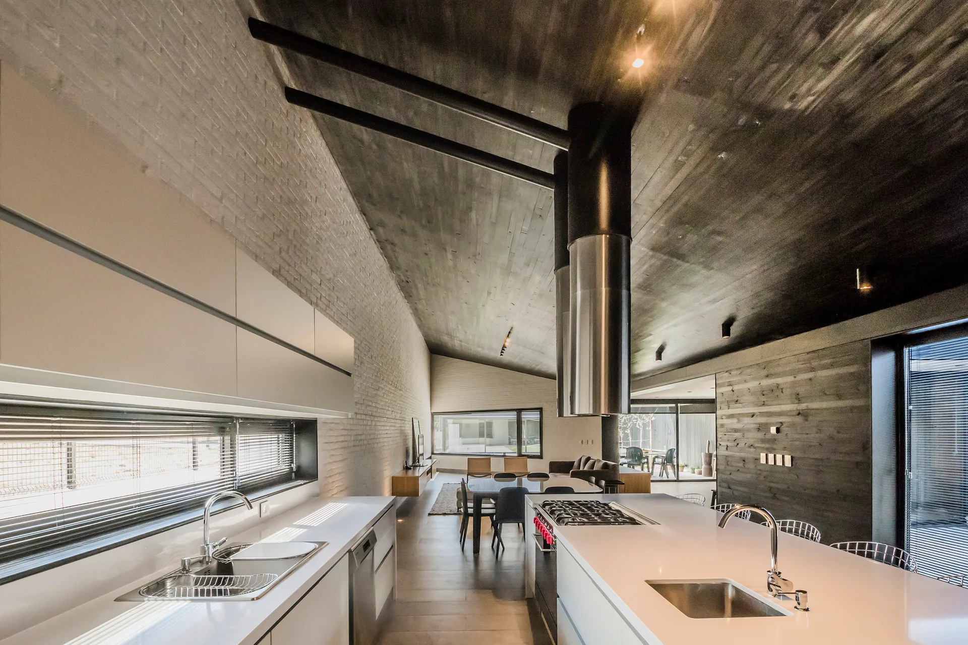 A modern open-concept kitchen featuring a long white island, a dark wood sloped ceiling, and a painted white brick wall.
