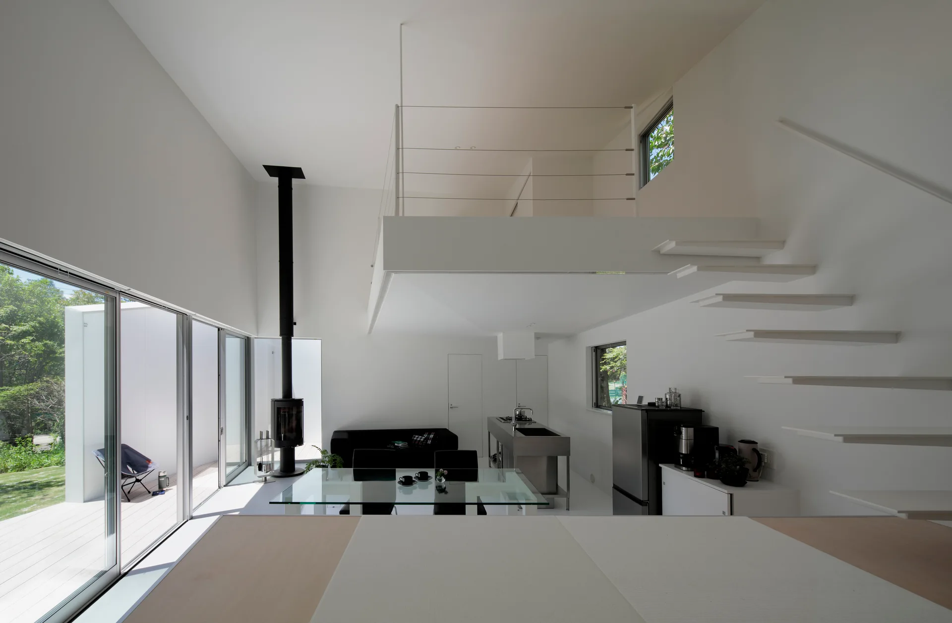 A minimalist white double-height living room featuring a floating staircase, mezzanine level, glass-top dining table, and tall black wood stove.