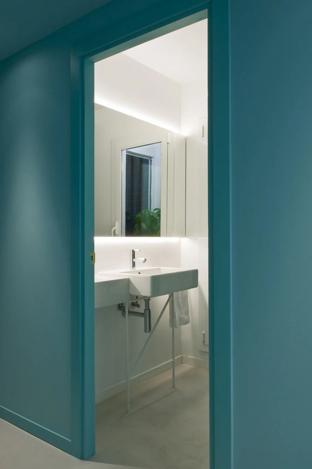 A view through a bright blue painted doorway into a minimalist white bathroom featuring a backlit rectangular mirror and a wall-mounted sink.