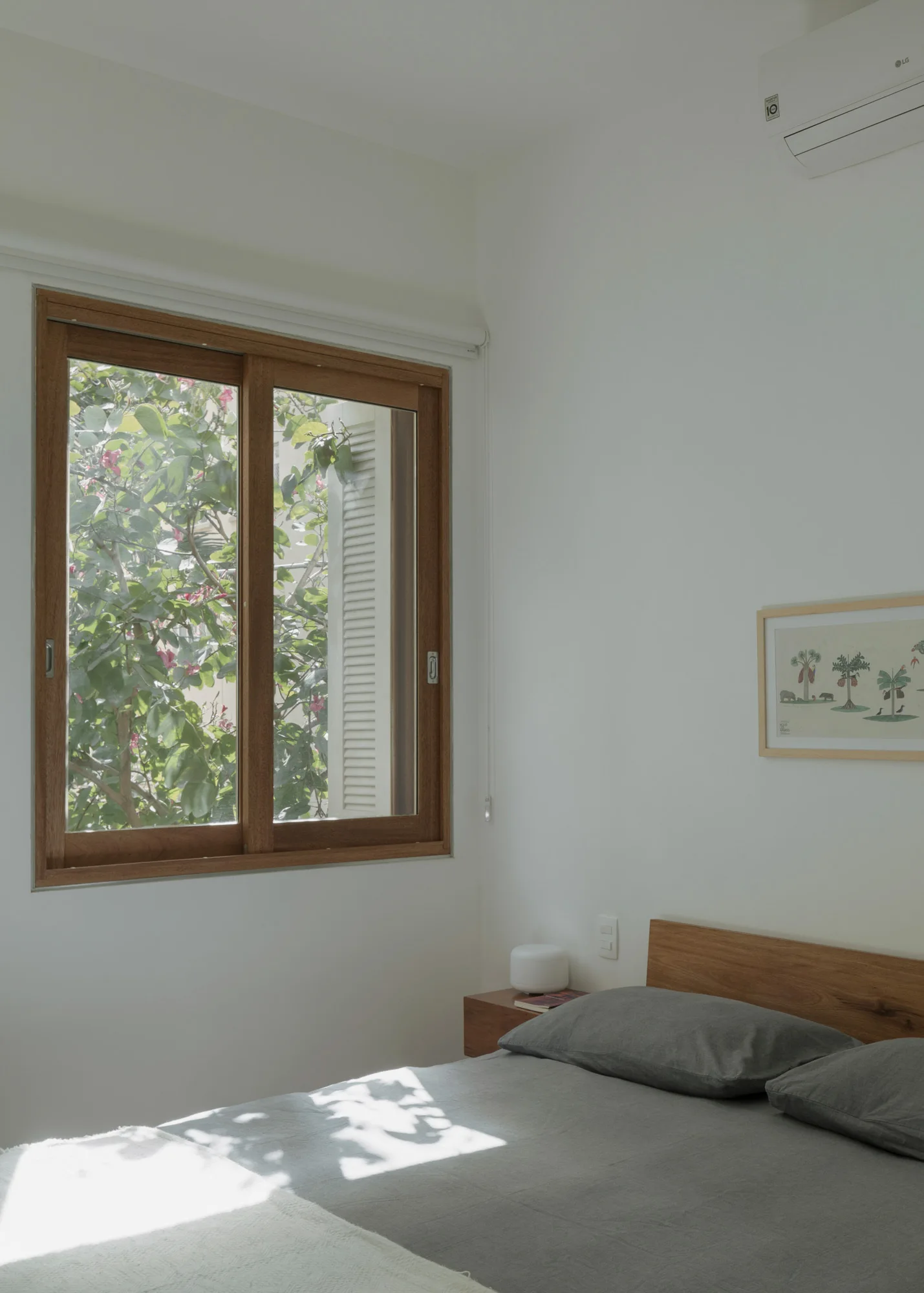 A minimalist bedroom with white walls, a wooden window looking out at green trees, and a bed with grey sheets and a wooden headboard.