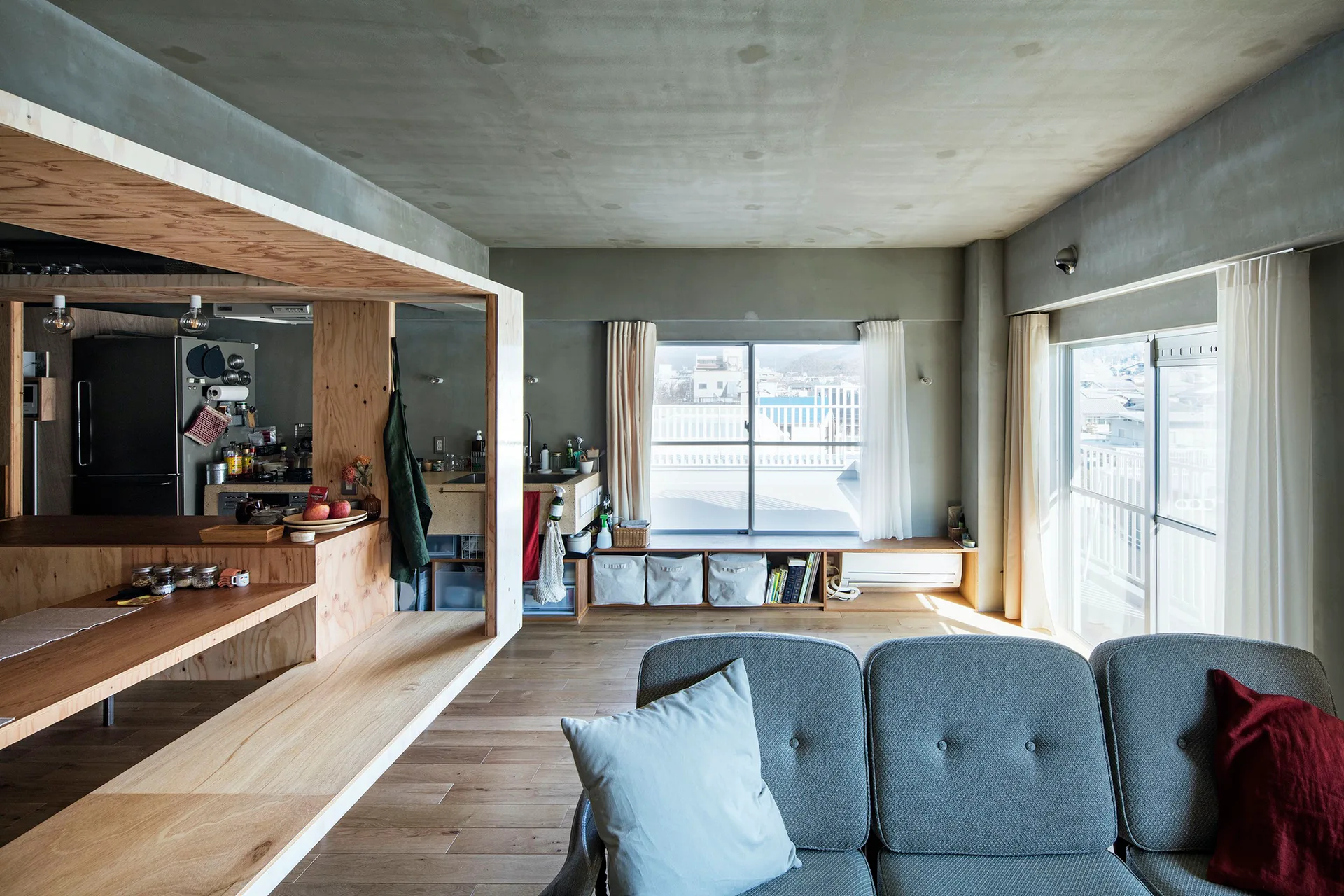 An open-plan living room with a grey tufted sofa, raw concrete walls, and a large custom plywood partition framing the kitchen.