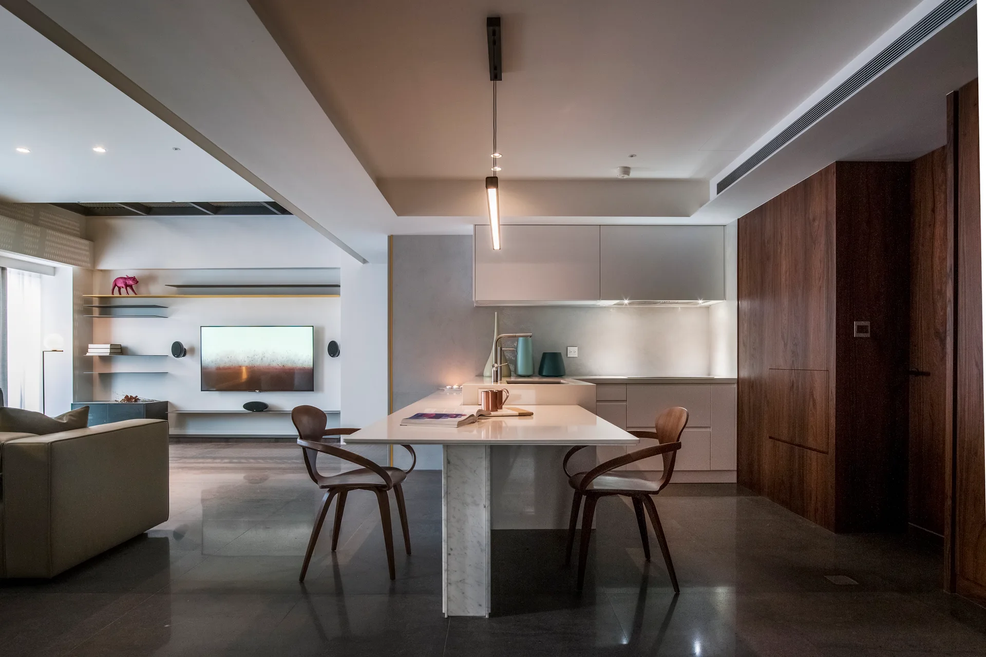 A modern kitchen island with a white marble leg and two curved wooden chairs sits adjacent to a wood-paneled wall and polished floor.