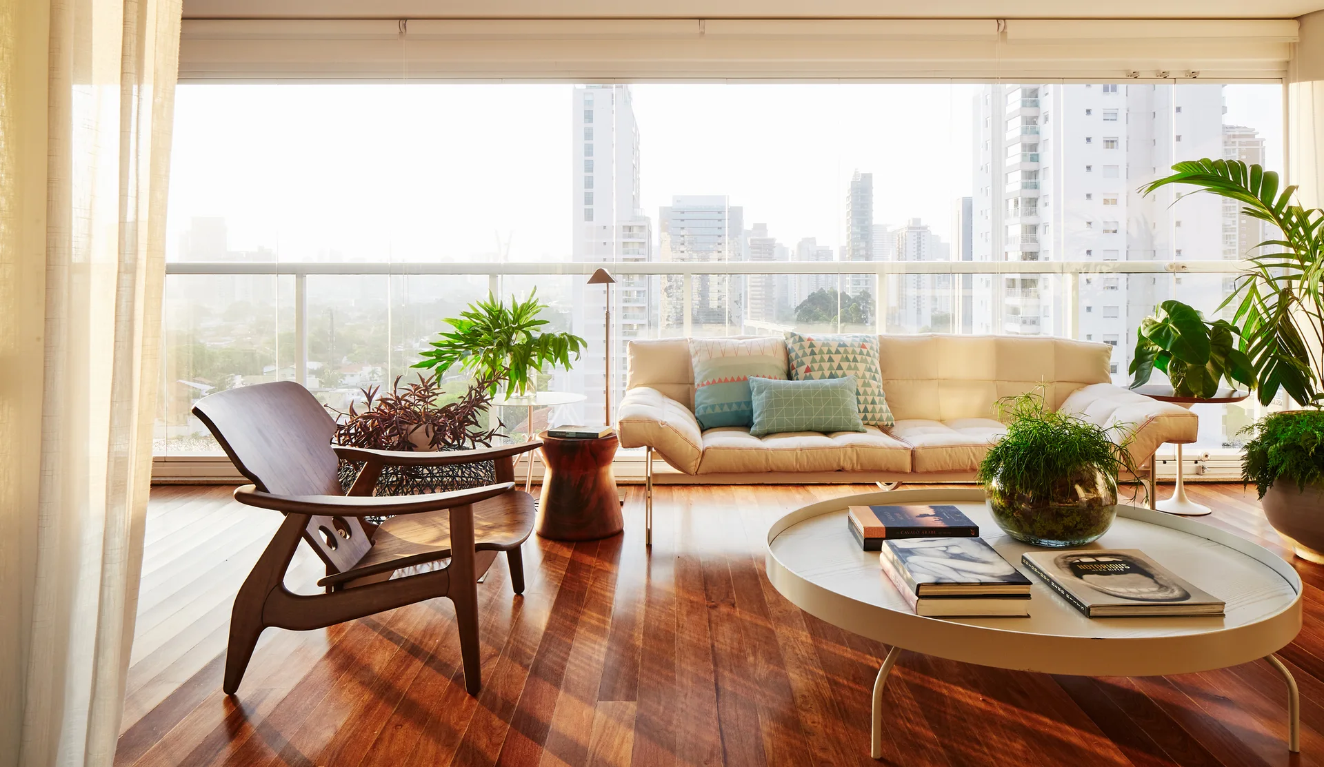 A sunlit living room features a beige sofa, a dark wood lounge chair, a low round coffee table, and large windows looking out at a city skyline.