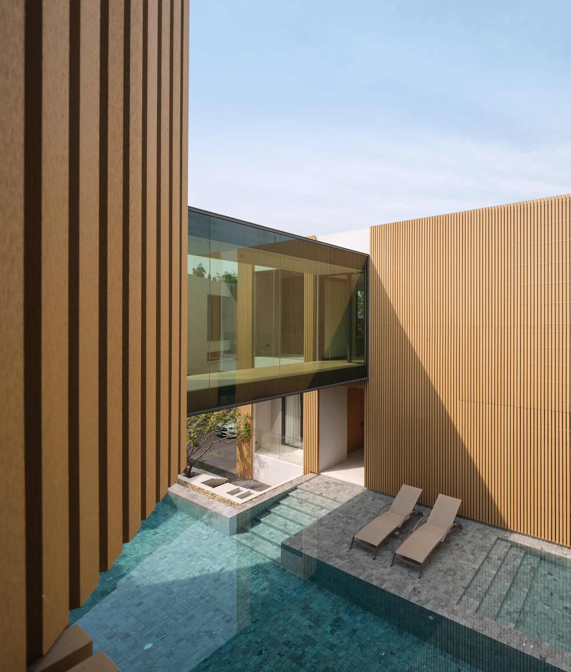 A modern courtyard pool area featuring a gray stone deck with two lounge chairs, vertical wood-slat walls, and an elevated glass walkway.