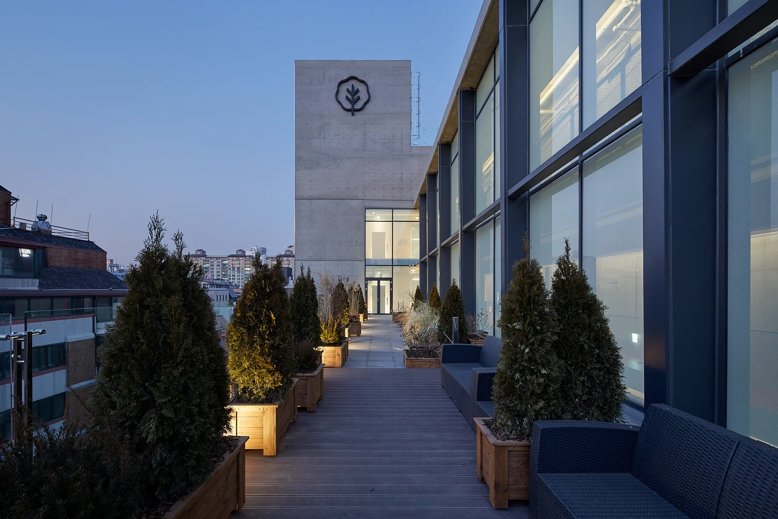 A modern rooftop terrace features a grey wooden deck, black wicker sofas, and a row of illuminated evergreen planters against a concrete building.