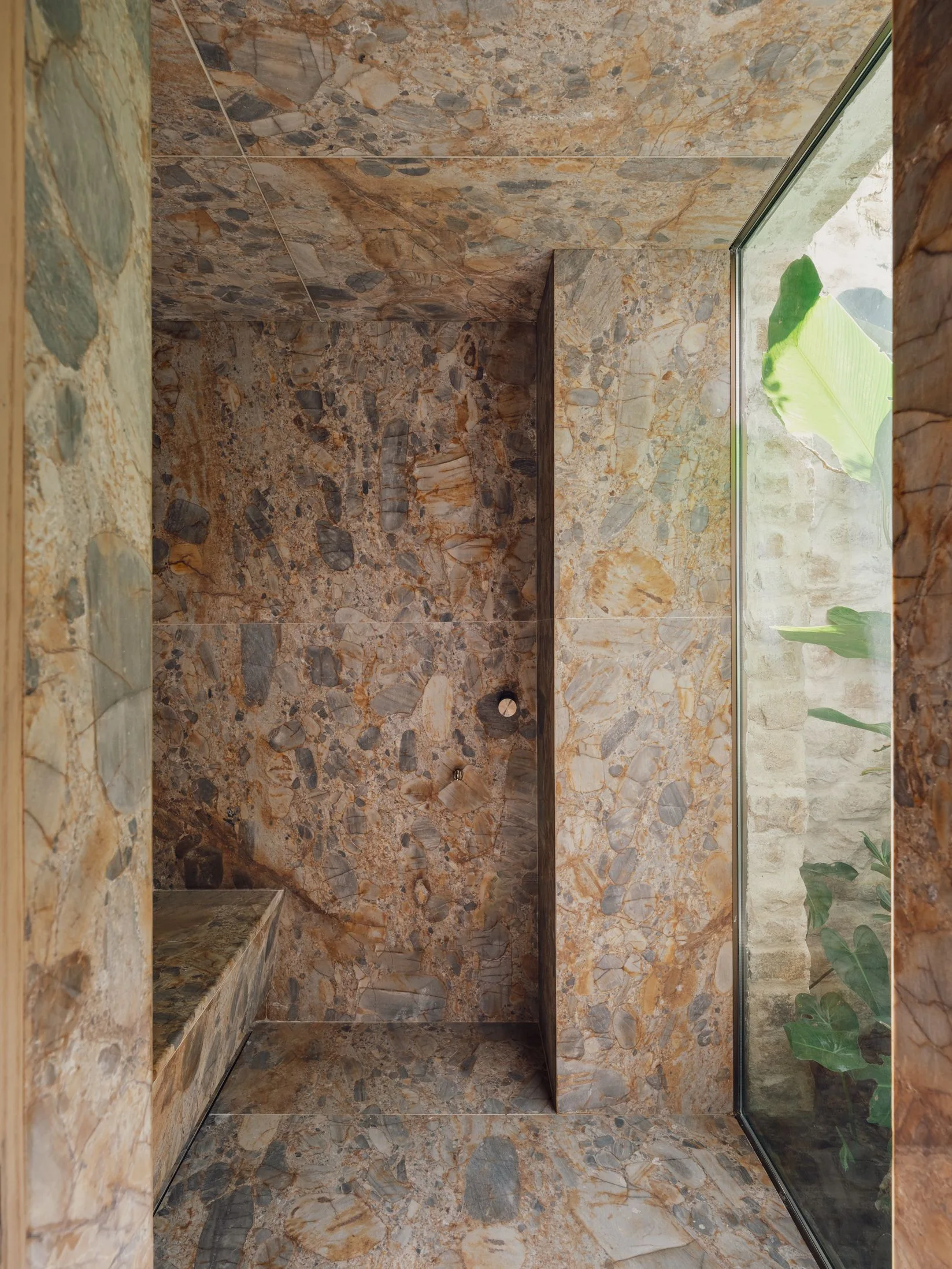 A walk-in shower clad entirely in variegated brown stone features a built-in bench and a tall glass window looking out at leafy green plants.