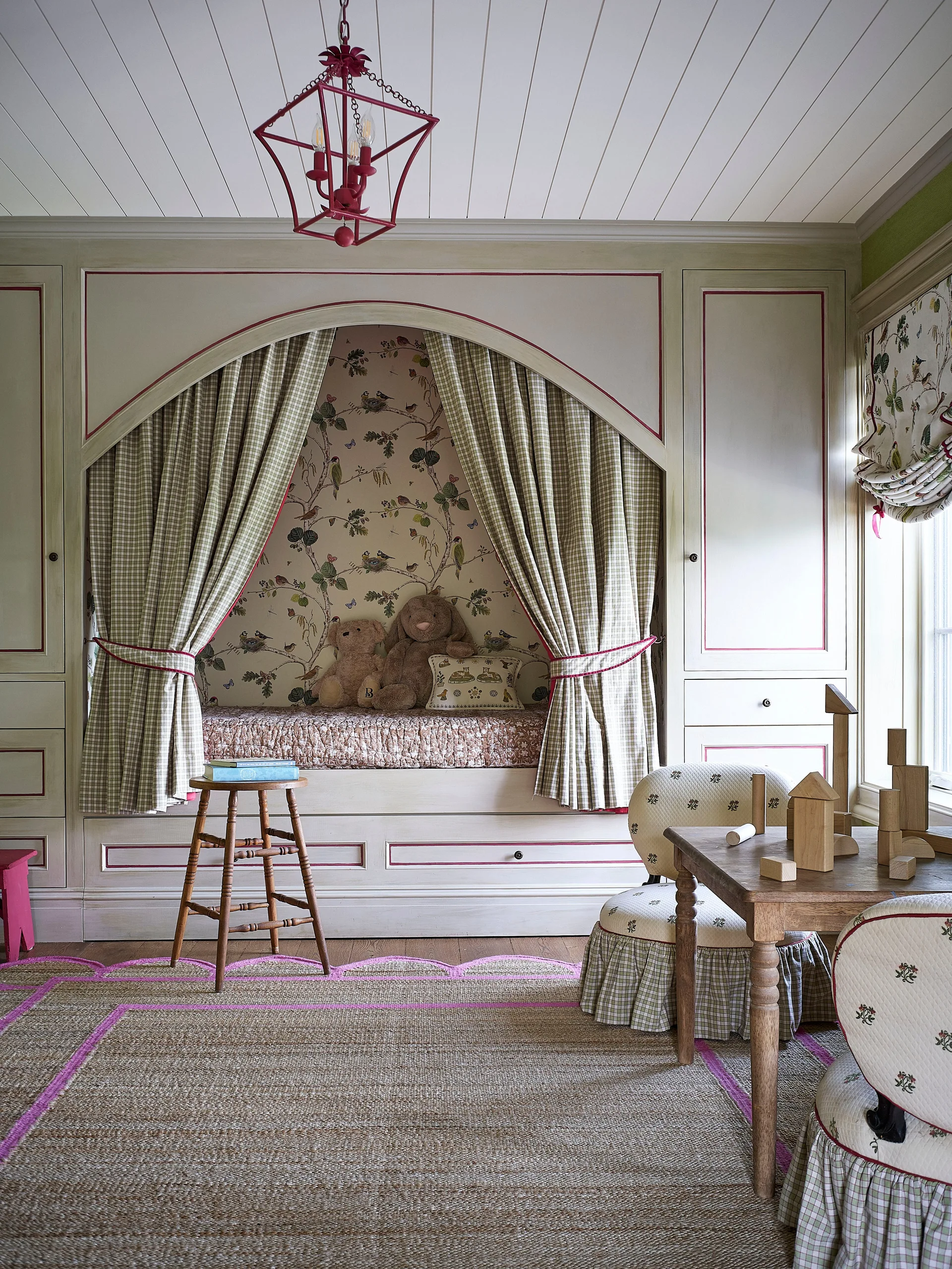 A children's playroom features a built-in daybed nook framed by green checkered curtains, botanical wallpaper, and a small wooden play table.