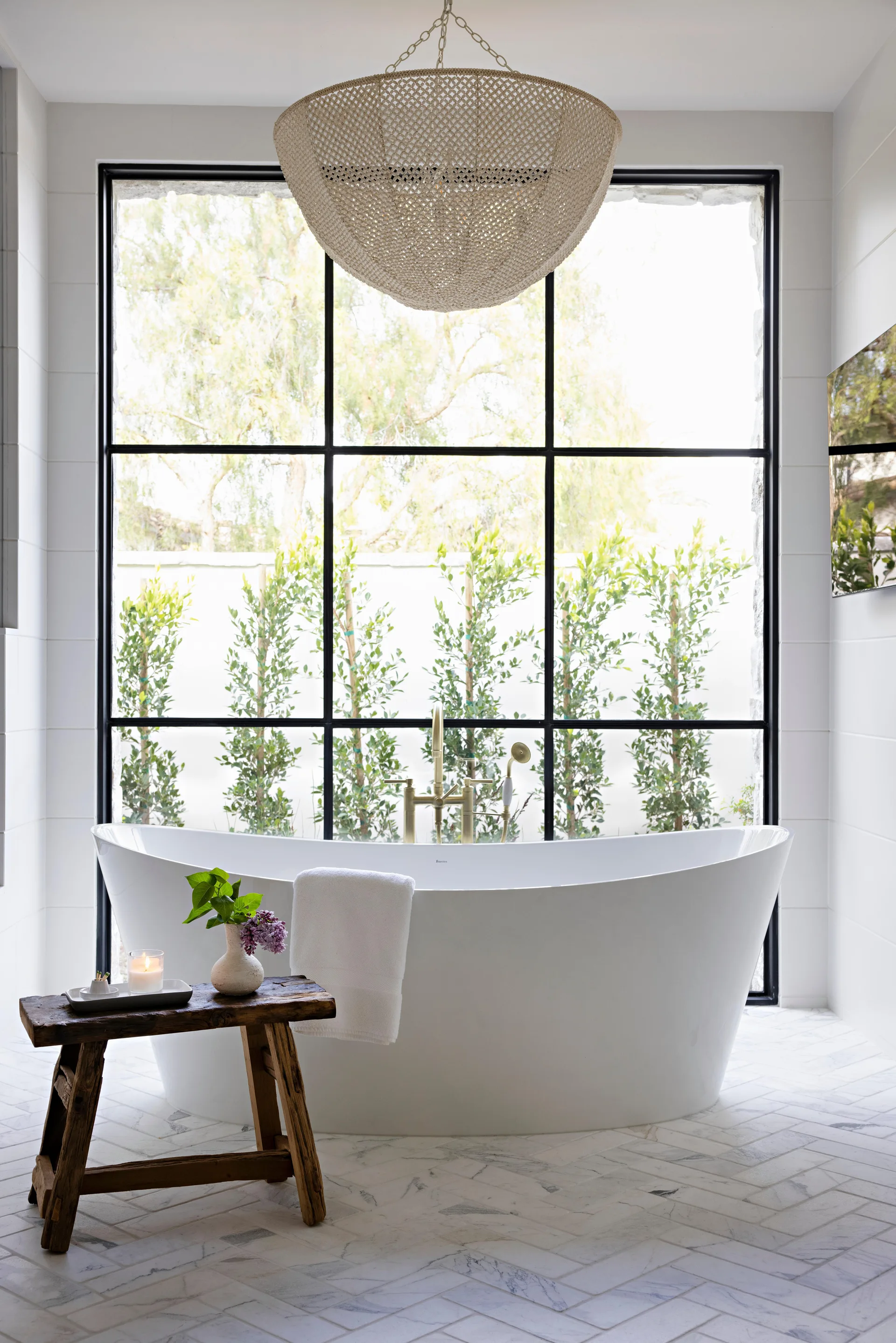 A white freestanding bathtub stands on herringbone marble tile floors in front of a large black-grid window with a woven pendant light above.