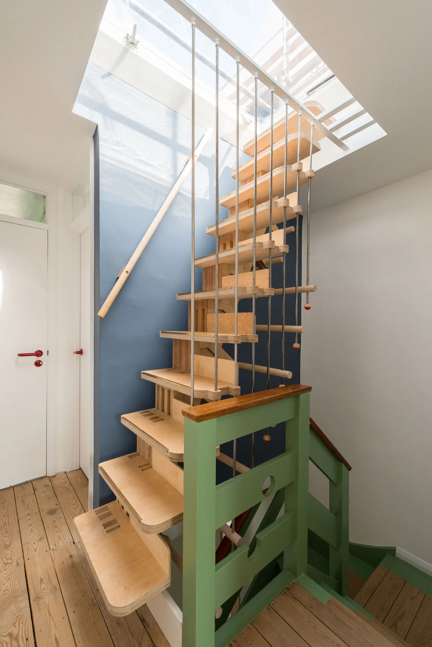 A custom plywood staircase with thin metal support rods rises toward an overhead skylight between a blue accent wall and white walls.