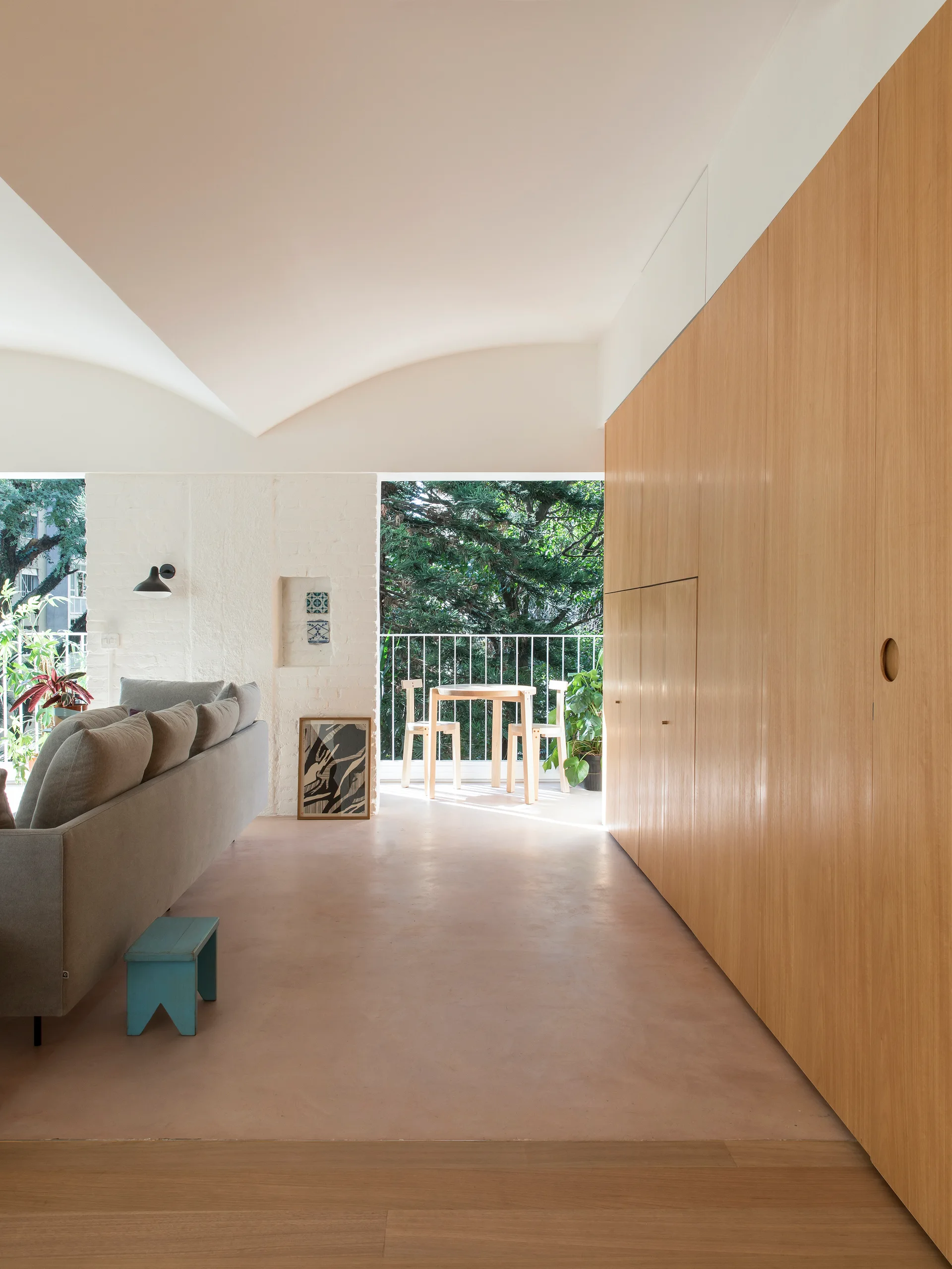 A minimalist living room with a grey sofa, vaulted ceiling, and built-in wood cabinetry opens onto a balcony with a small table and chairs.