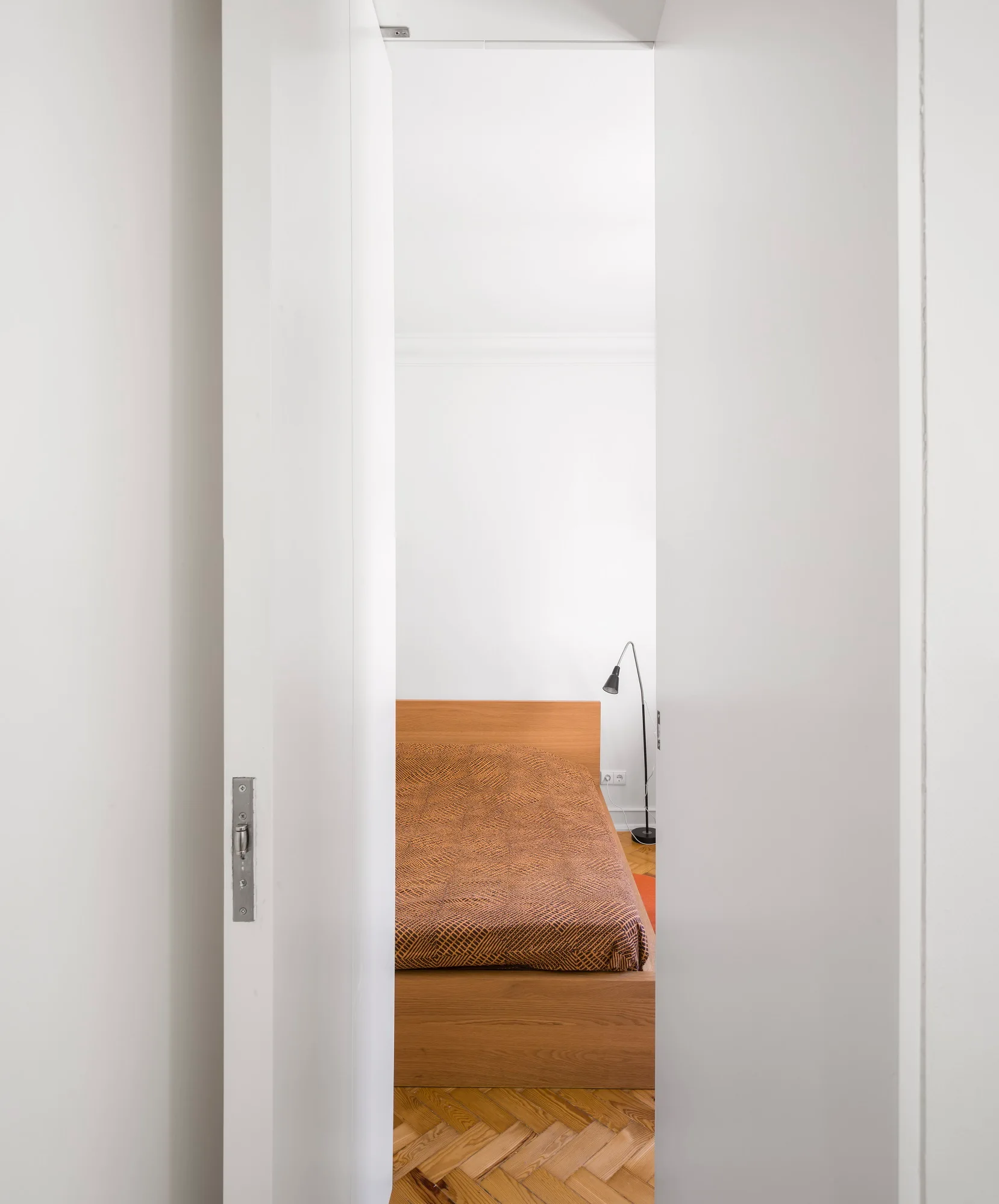 A view through a white doorway reveals a minimalist bedroom with a wooden bed frame, orange patterned bedding, and herringbone parquet flooring.