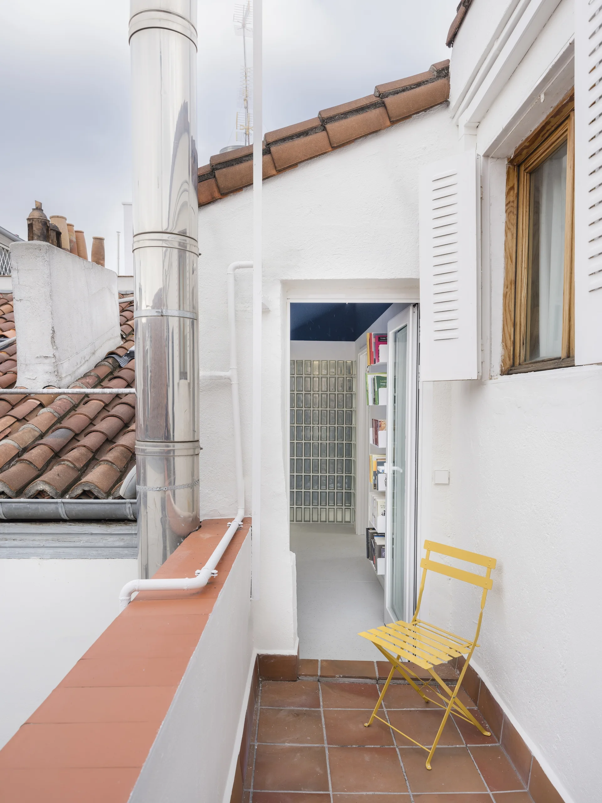 An outdoor rooftop balcony features terracotta tile flooring, white stucco walls, a yellow metal folding chair, and an open doorway showing a glass block wall.