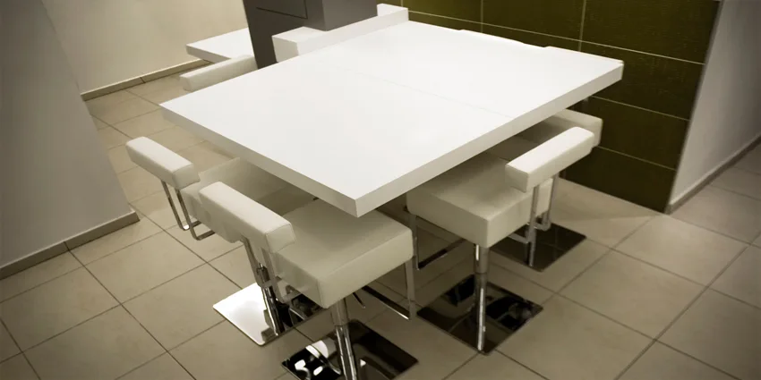 A modern white dining table surrounded by upholstered white swivel bar stools with chrome bases in a kitchen with olive green wall tiles.