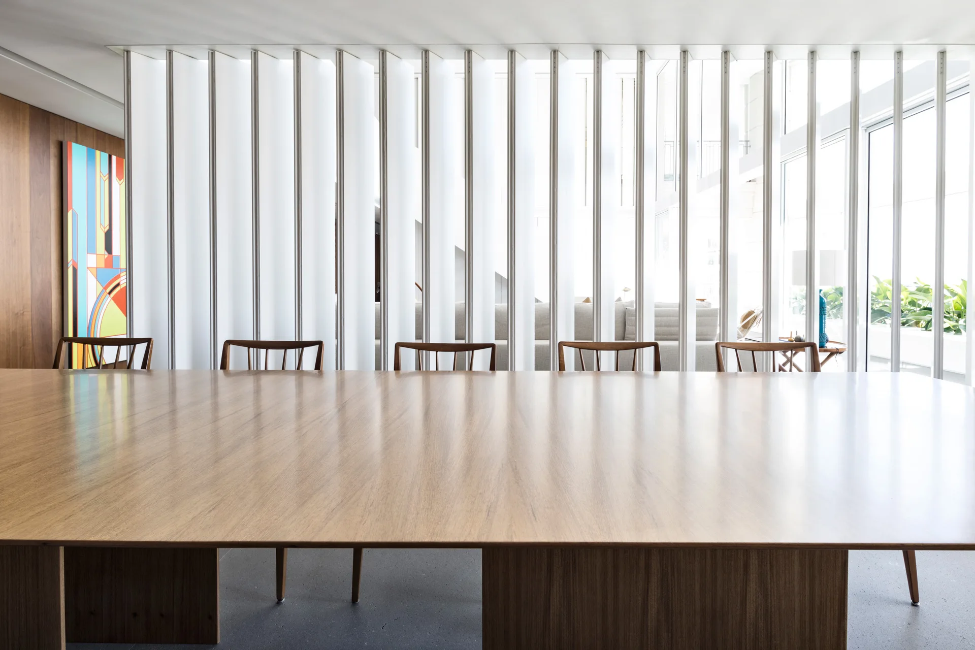 A long wooden dining table and chairs stand before a floor-to-ceiling white slat partition screen in a bright minimalist room.