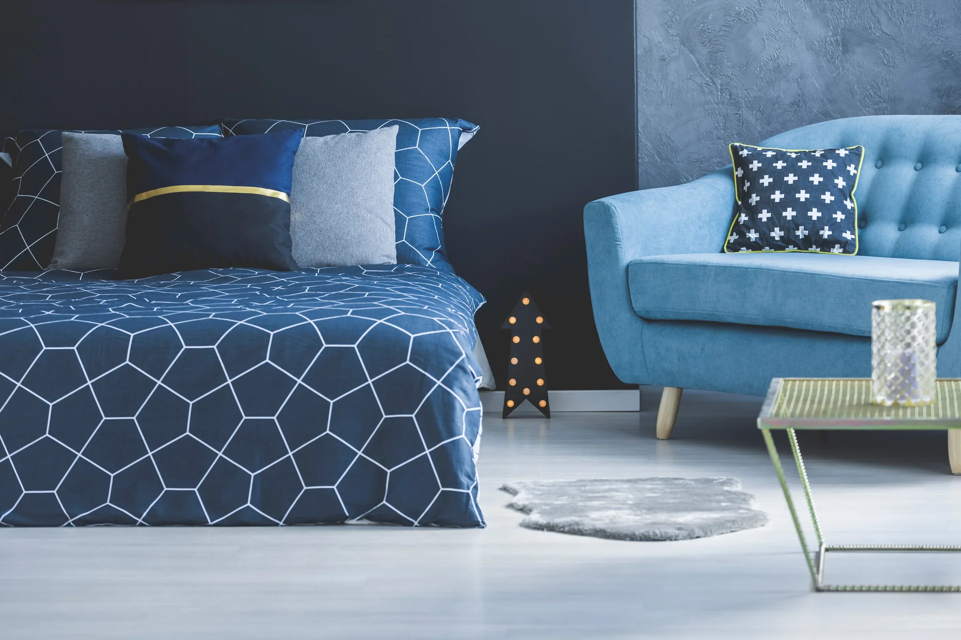 A modern bedroom features a bed with blue geometric patterned bedding next to a blue tufted armchair and a gold metal side table on light floors.