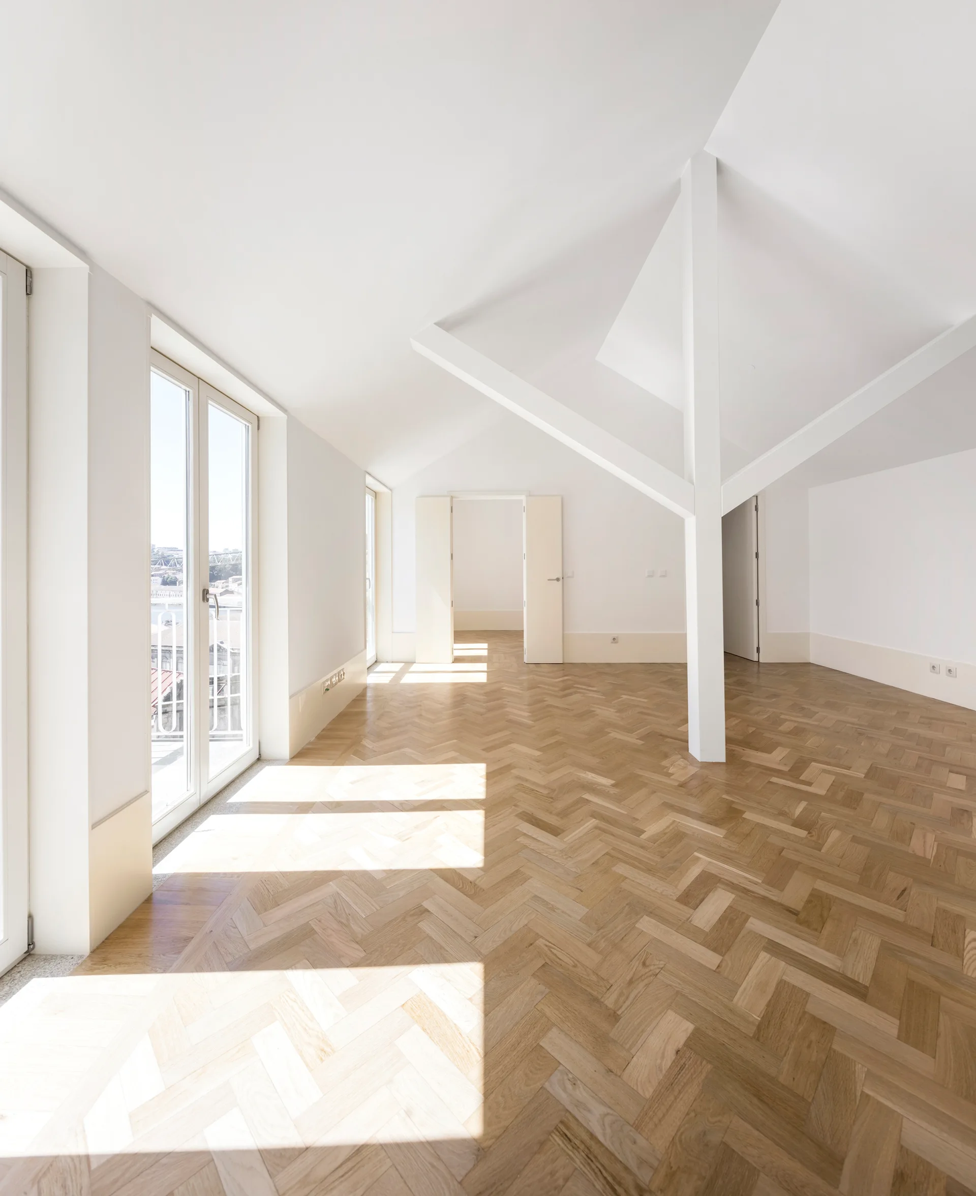 An empty loft space with white sloped ceilings, a central painted support beam, and light brown herringbone parquet wood flooring.