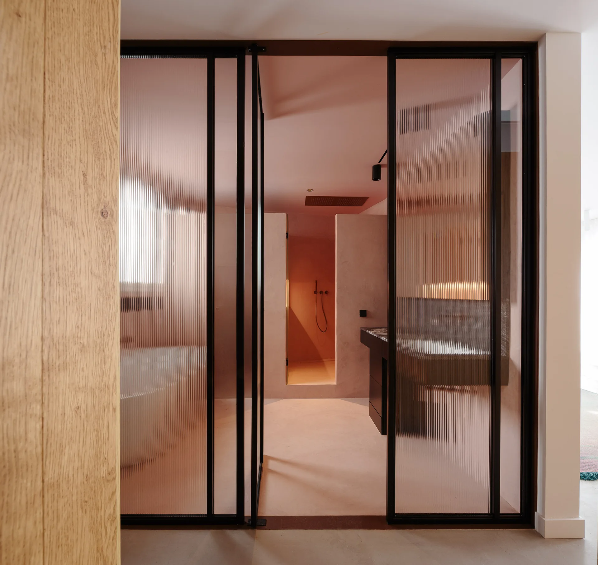 A modern monochromatic pink bathroom is framed by floor-to-ceiling black metal sliding doors with ribbed glass panels.