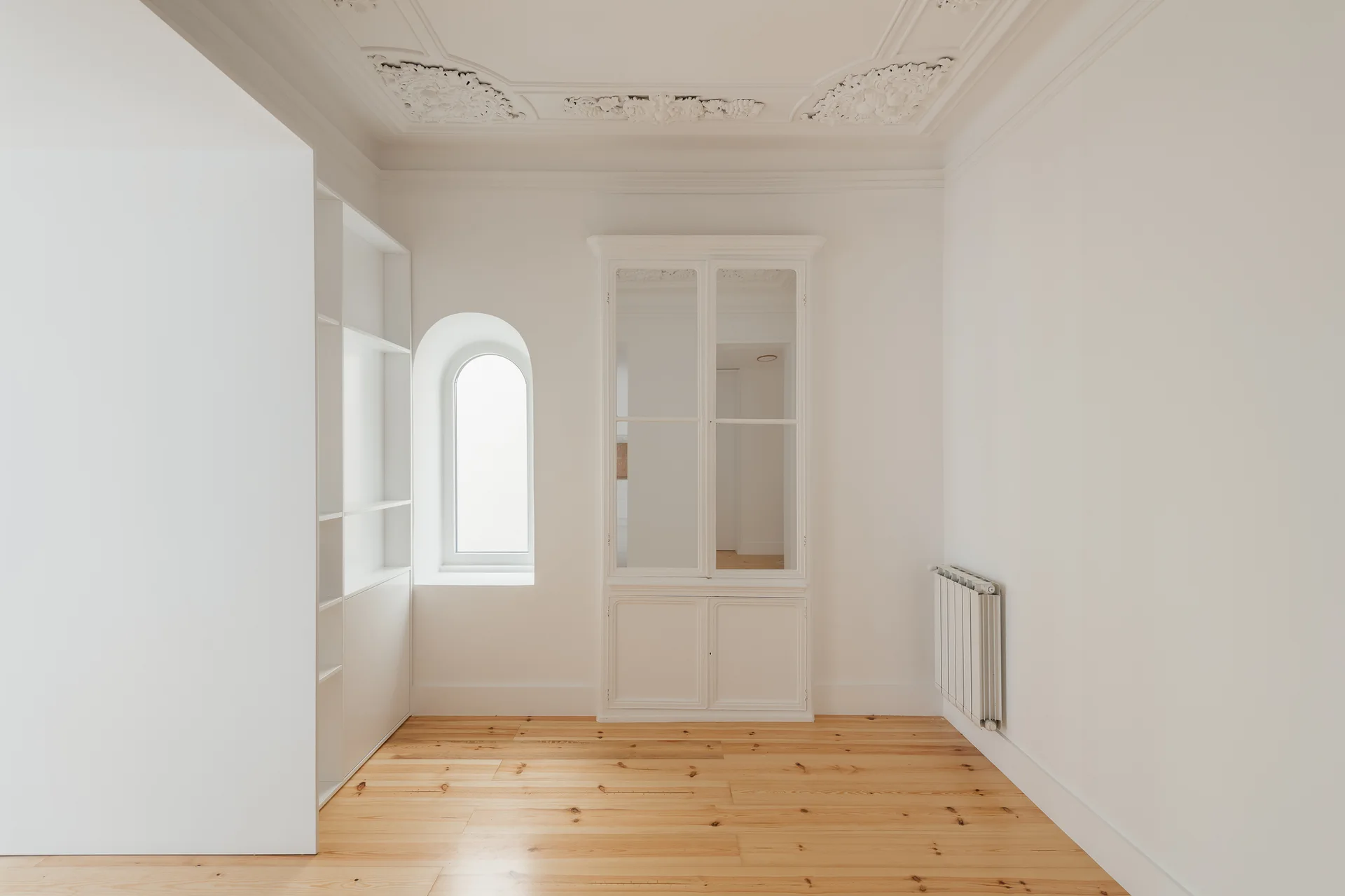 An empty white room with light hardwood floors, a built-in glass-front cabinet, an arched window, and ornate plaster ceiling molding.