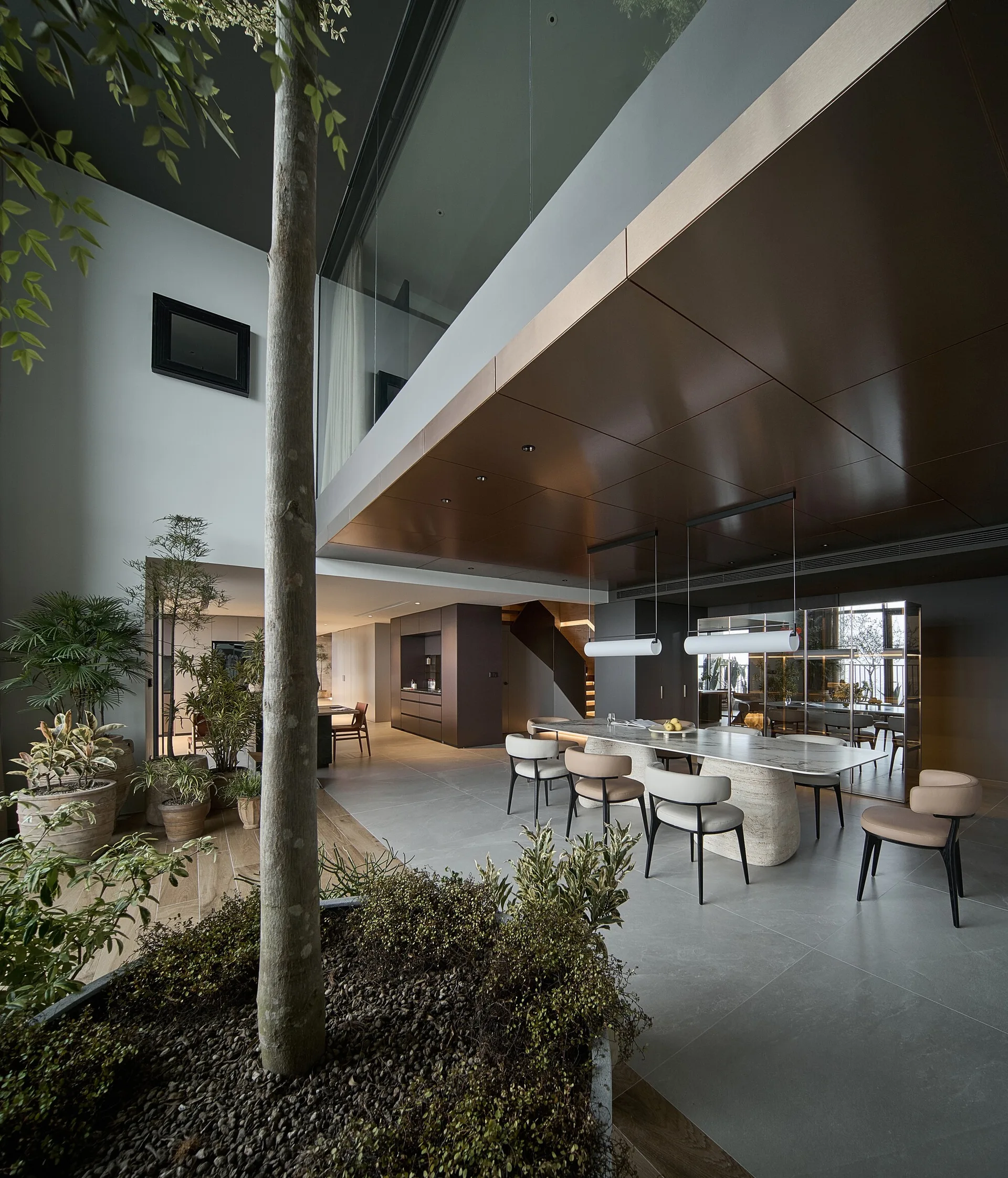 A spacious double-height dining room features a large stone table with beige chairs, a dark bronze ceiling panel, and a live tree planted indoors.
