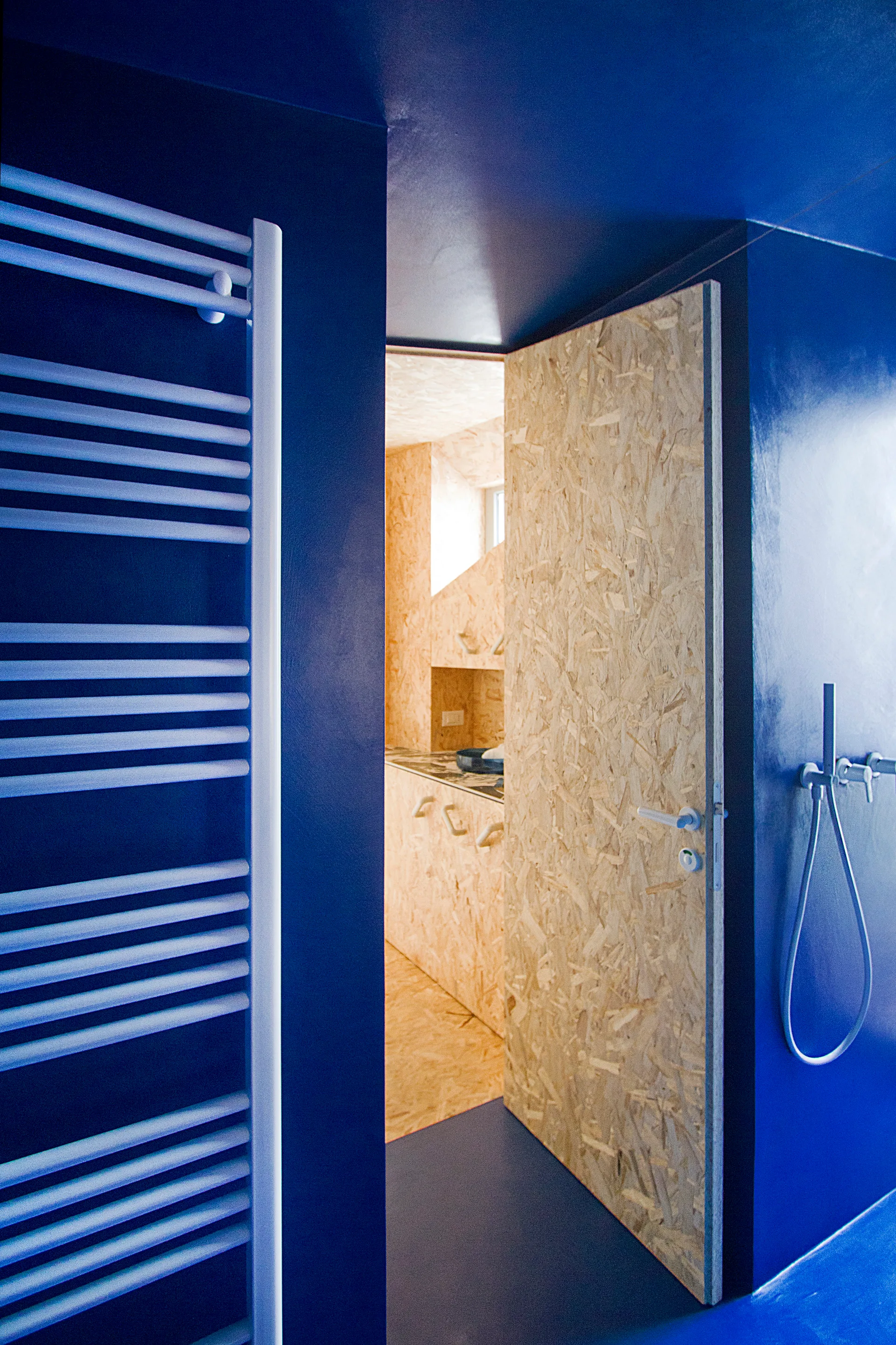 A vibrant blue bathroom with a white ladder towel warmer features an open door made of textured oriented strand board.
