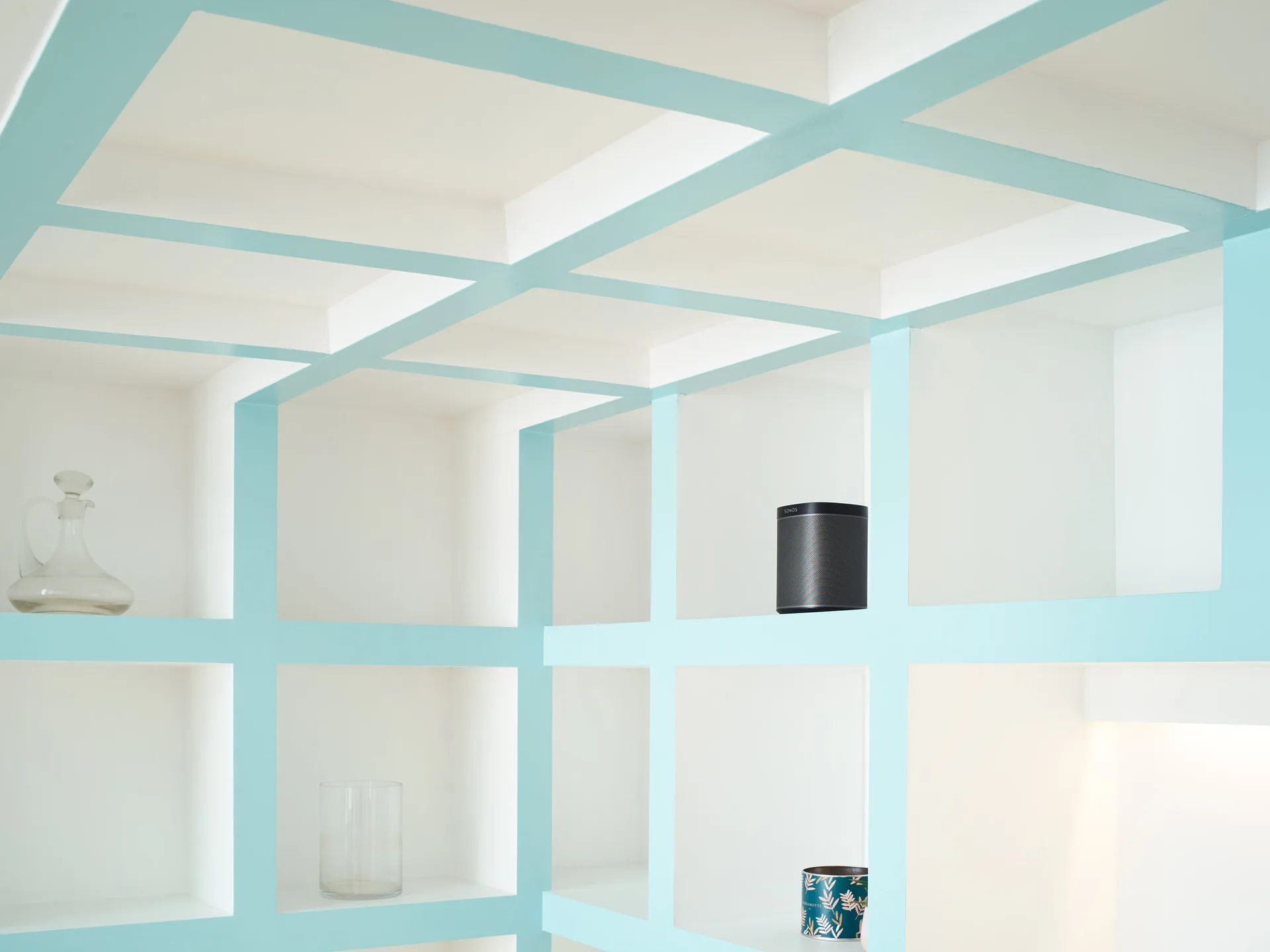 A close-up of custom white grid shelving with light blue painted trim, holding a glass decanter and a small speaker.