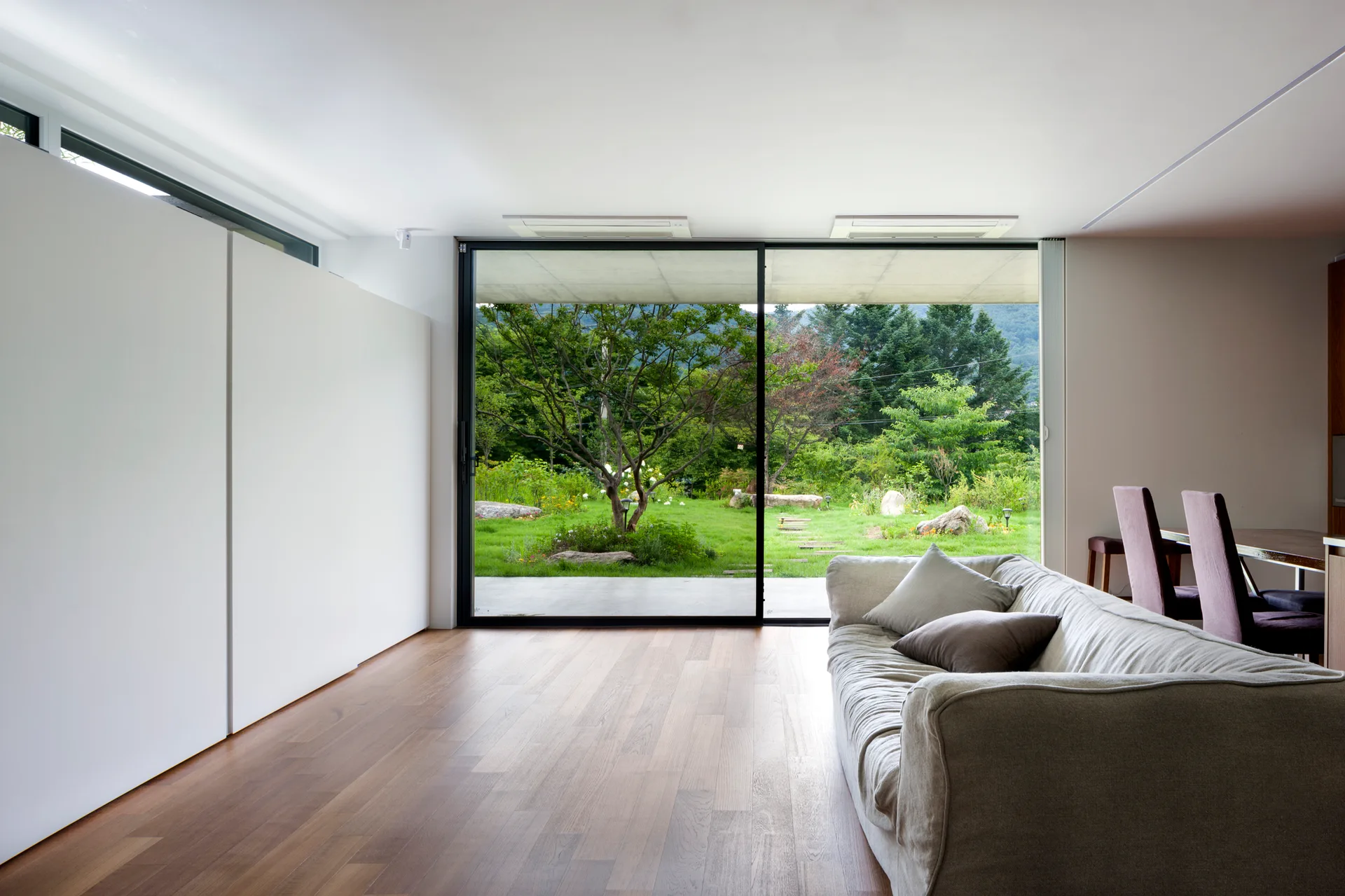 A minimalist living room features a beige fabric sofa on wood floors next to large sliding glass doors showing a green lawn and trees.