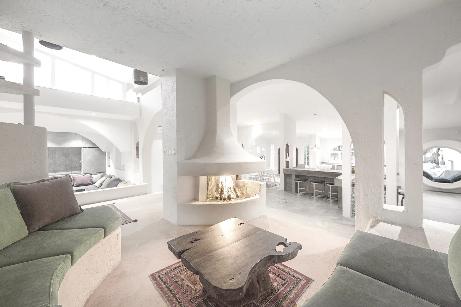 A spacious white stucco living room with a central hooded fireplace, built-in green sofas, and a rustic wood slab coffee table.