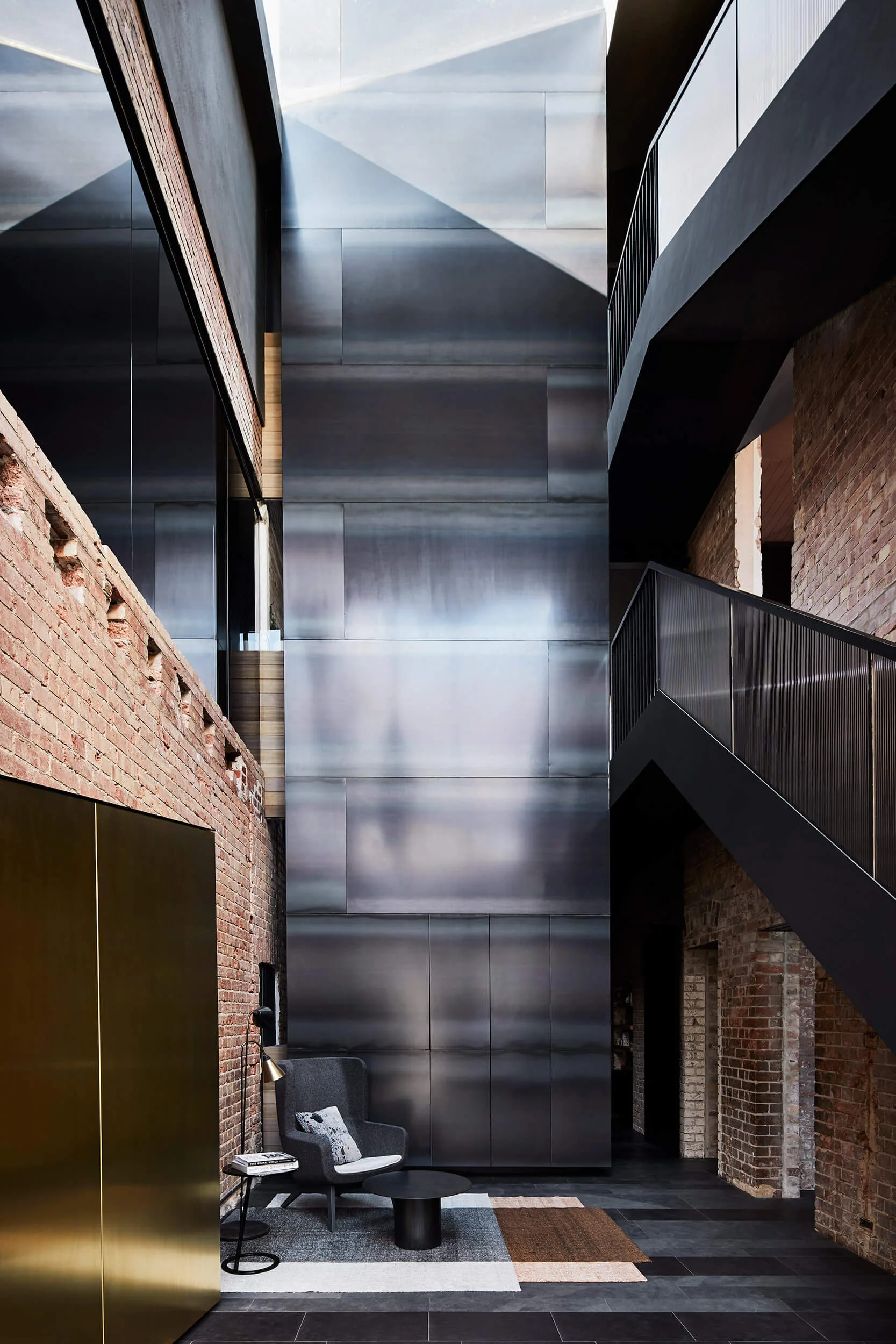 A multi-level modern atrium featuring a black metal staircase, raw exposed brick walls, and dark steel paneling above a small seating area.