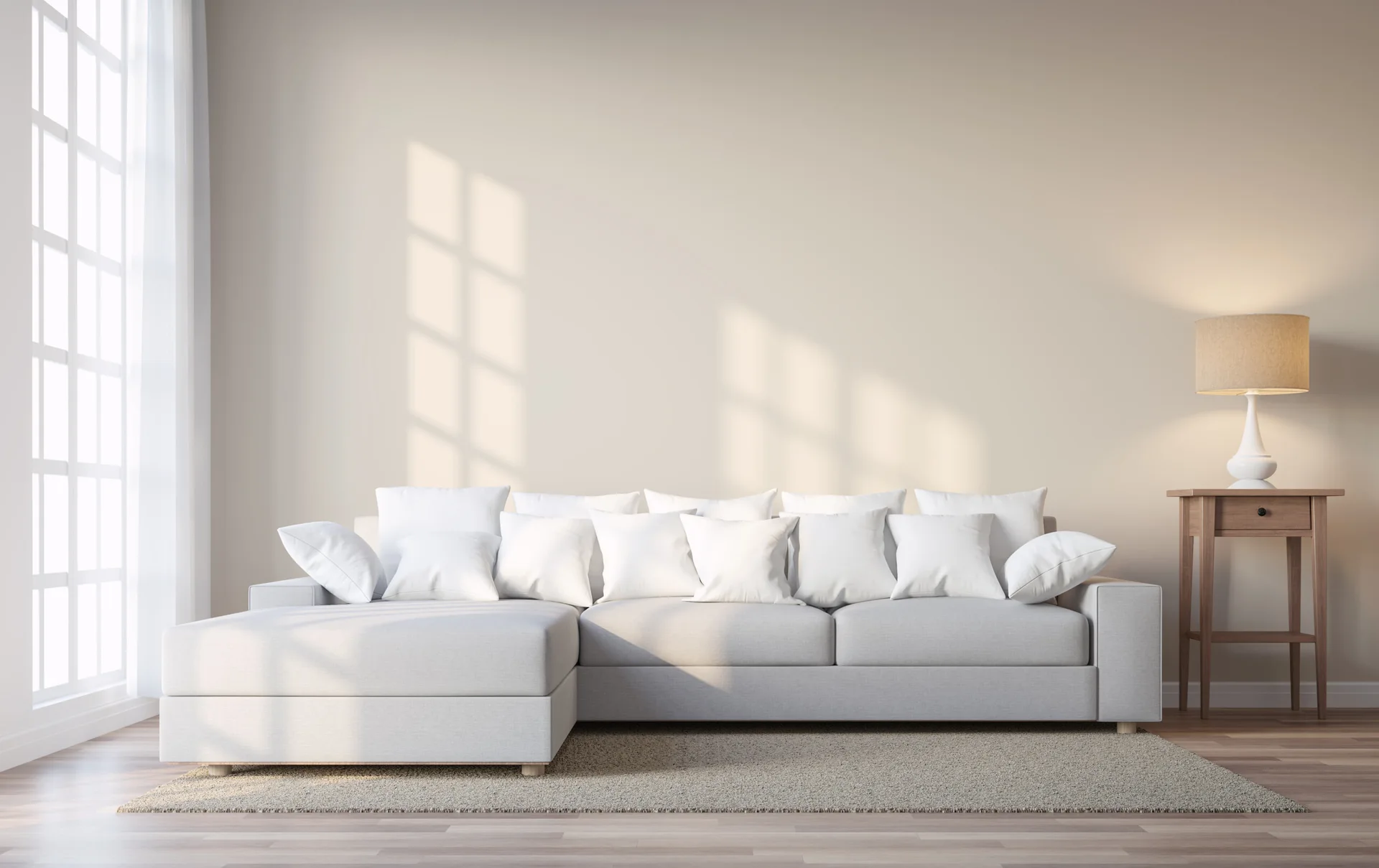 A light gray sectional sofa with white throw pillows sits against a beige wall next to a wooden side table with a lit lamp.