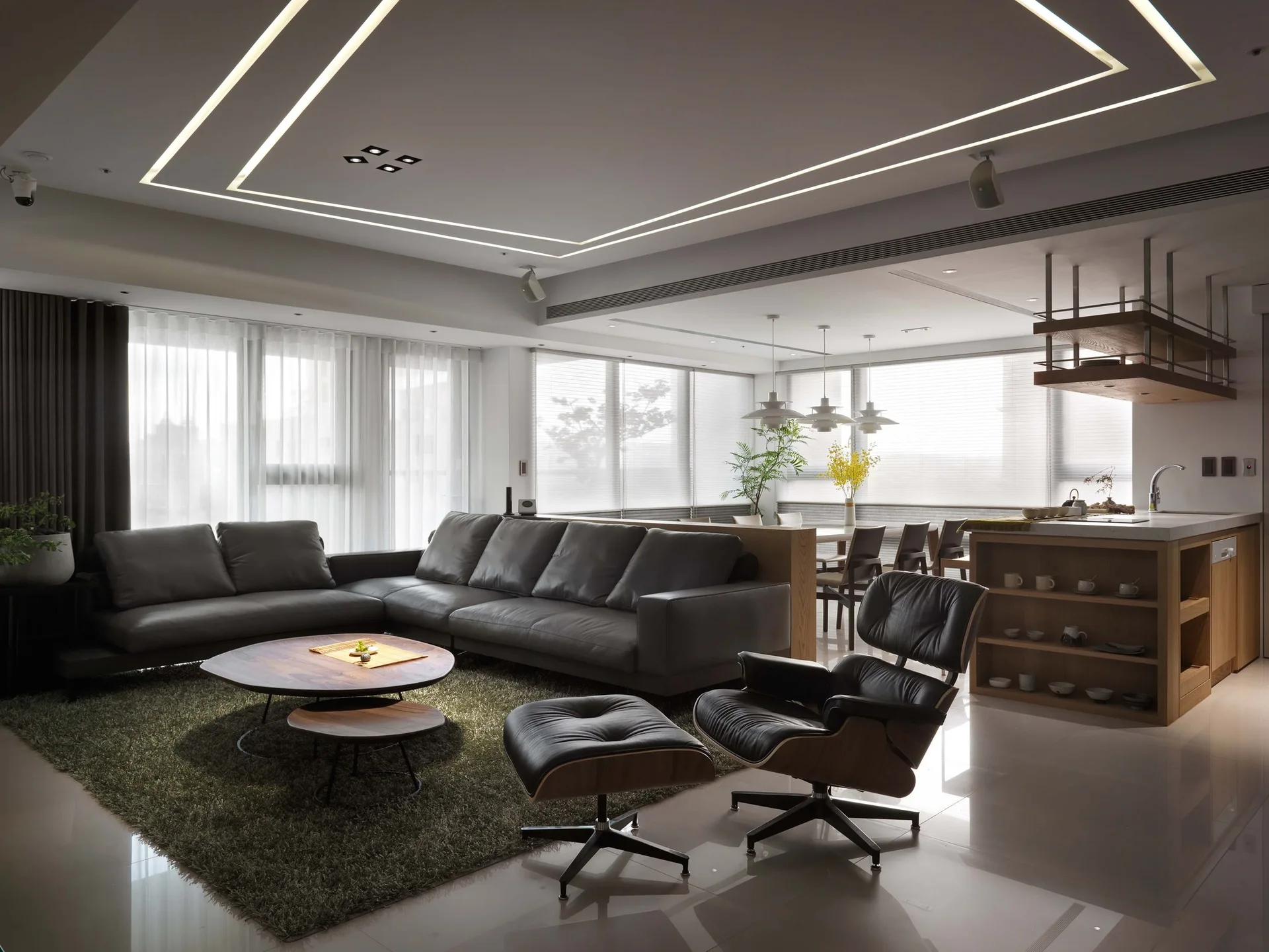 An open plan living room featuring a large grey sectional sofa, classic black leather lounge chair, and recessed linear ceiling light strips.