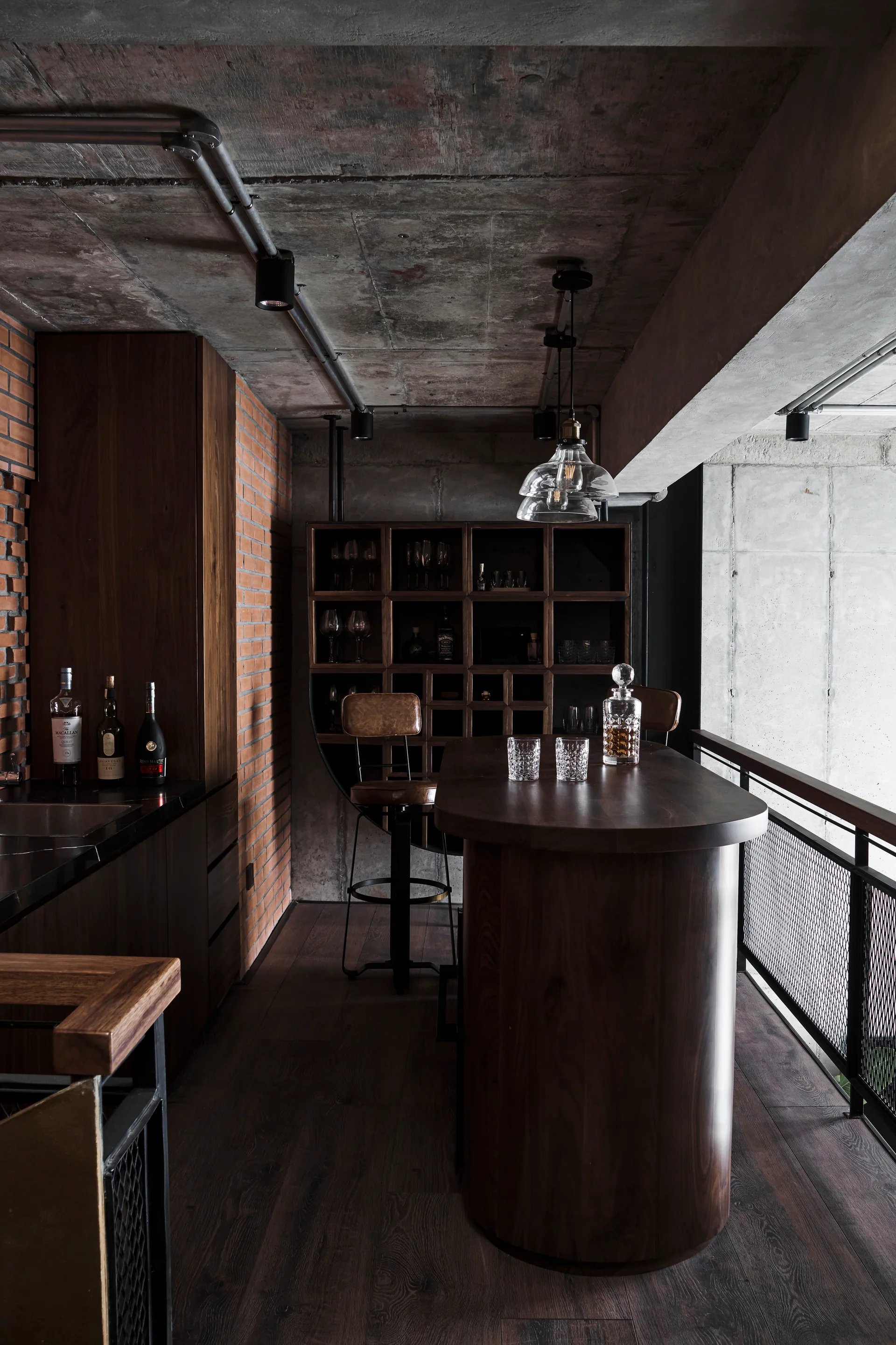 An industrial home bar features a curved dark wood counter, leather bar stools, an exposed concrete ceiling, and a red brick wall.