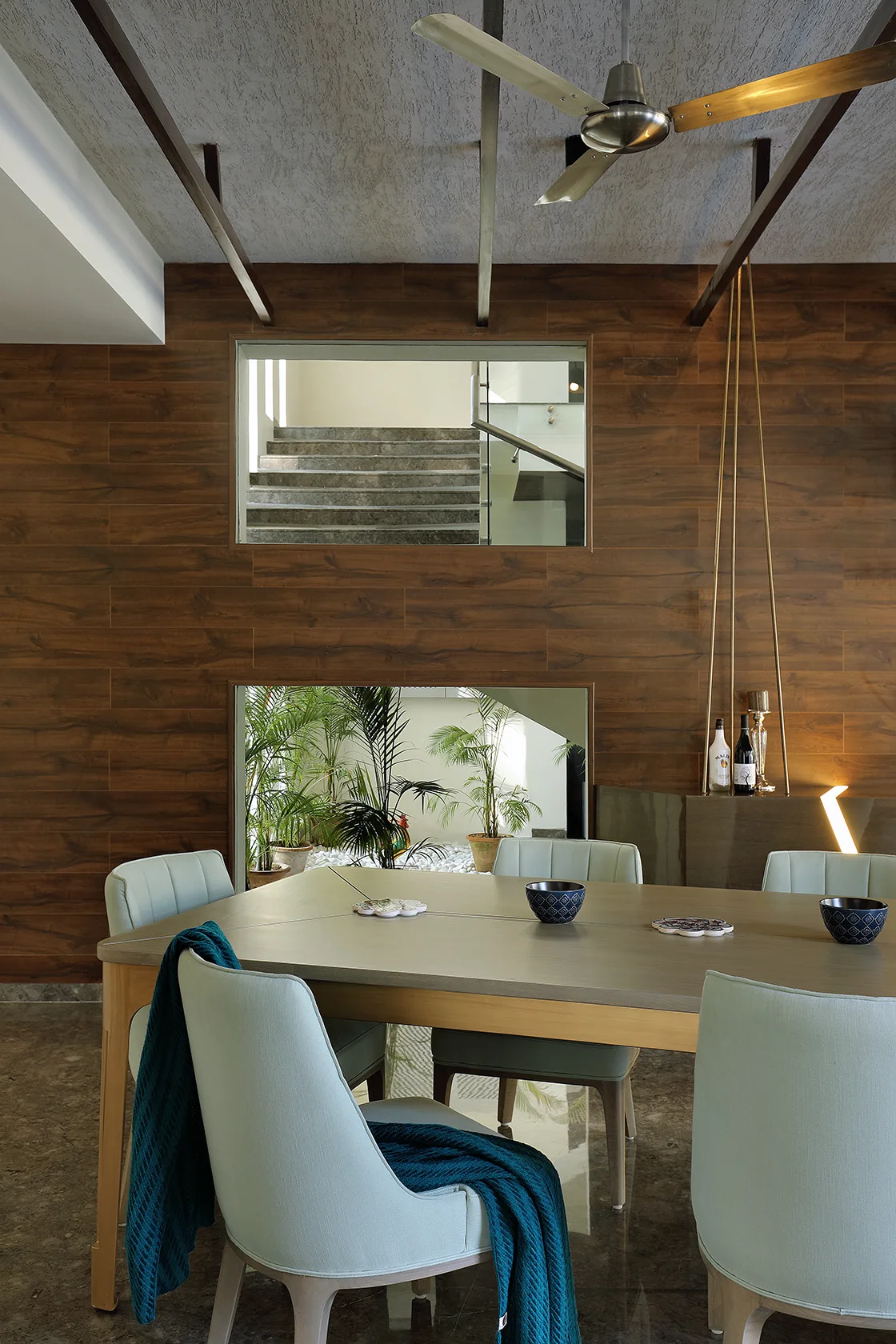 A modern dining room featuring a wooden dining table, mint green upholstered chairs, and a wood-paneled wall with two interior window cutouts.