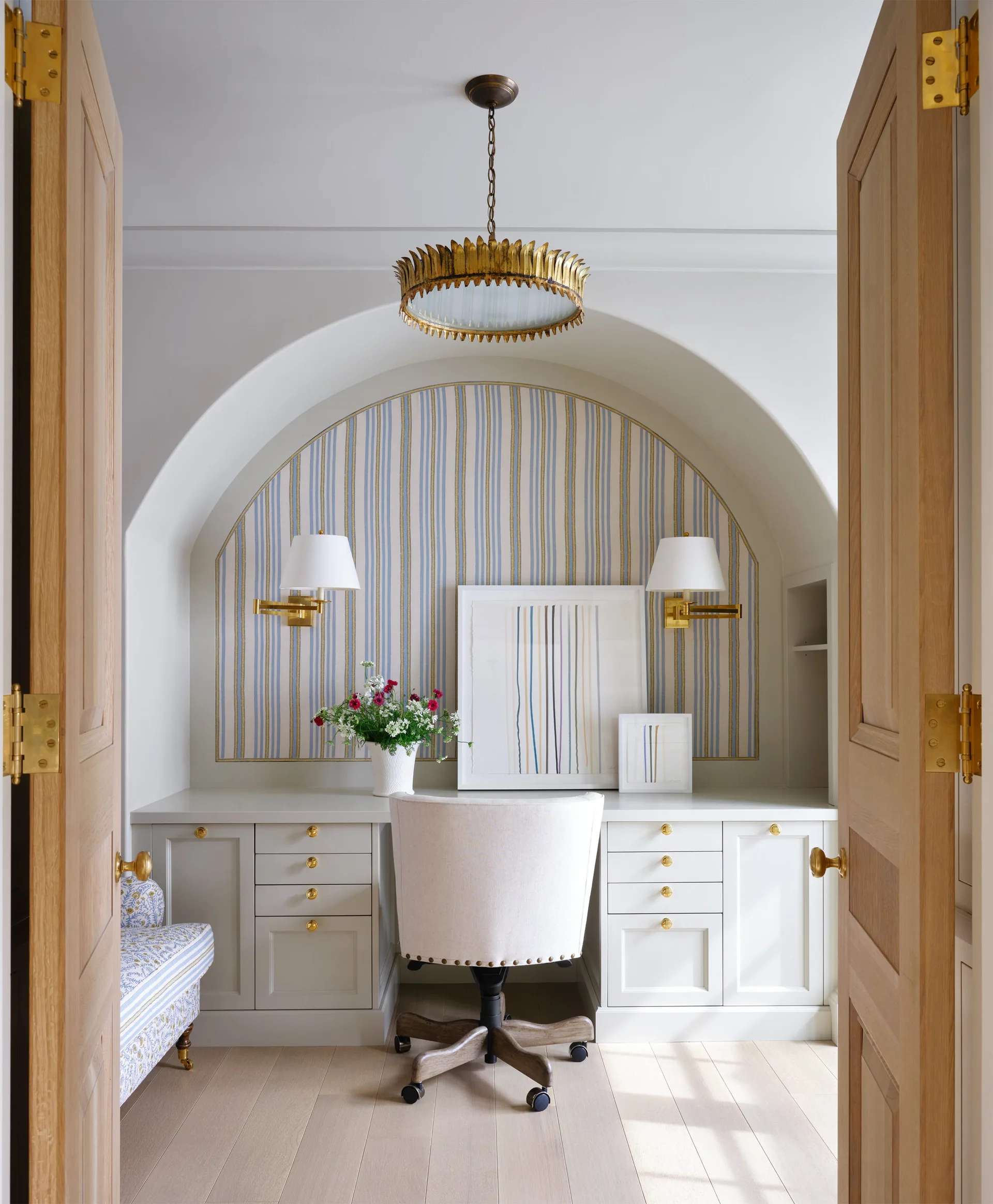 A home office desk built into an arched alcove featuring striped wallpaper, brass sconces, and a white upholstered desk chair on light wood floors.