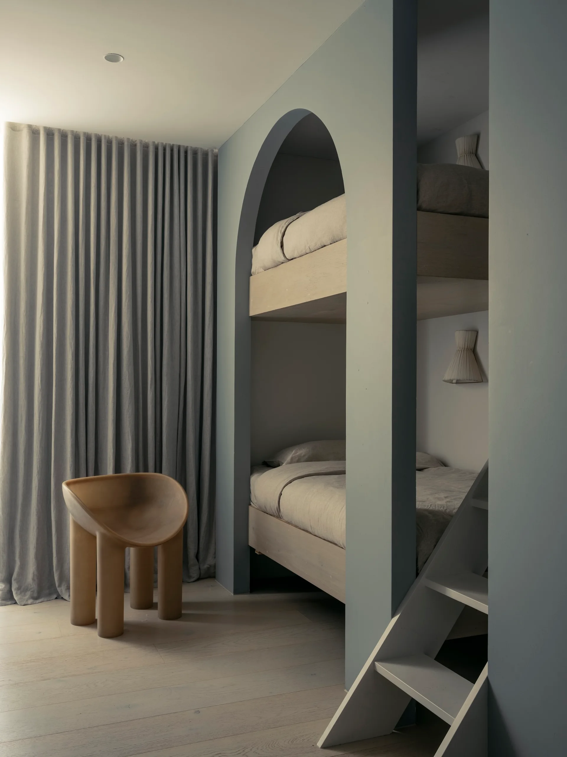 A minimalist kids bedroom featuring a blue built-in bunk bed with an arched opening, a sculptural tan chair, and grey floor-to-ceiling curtains.
