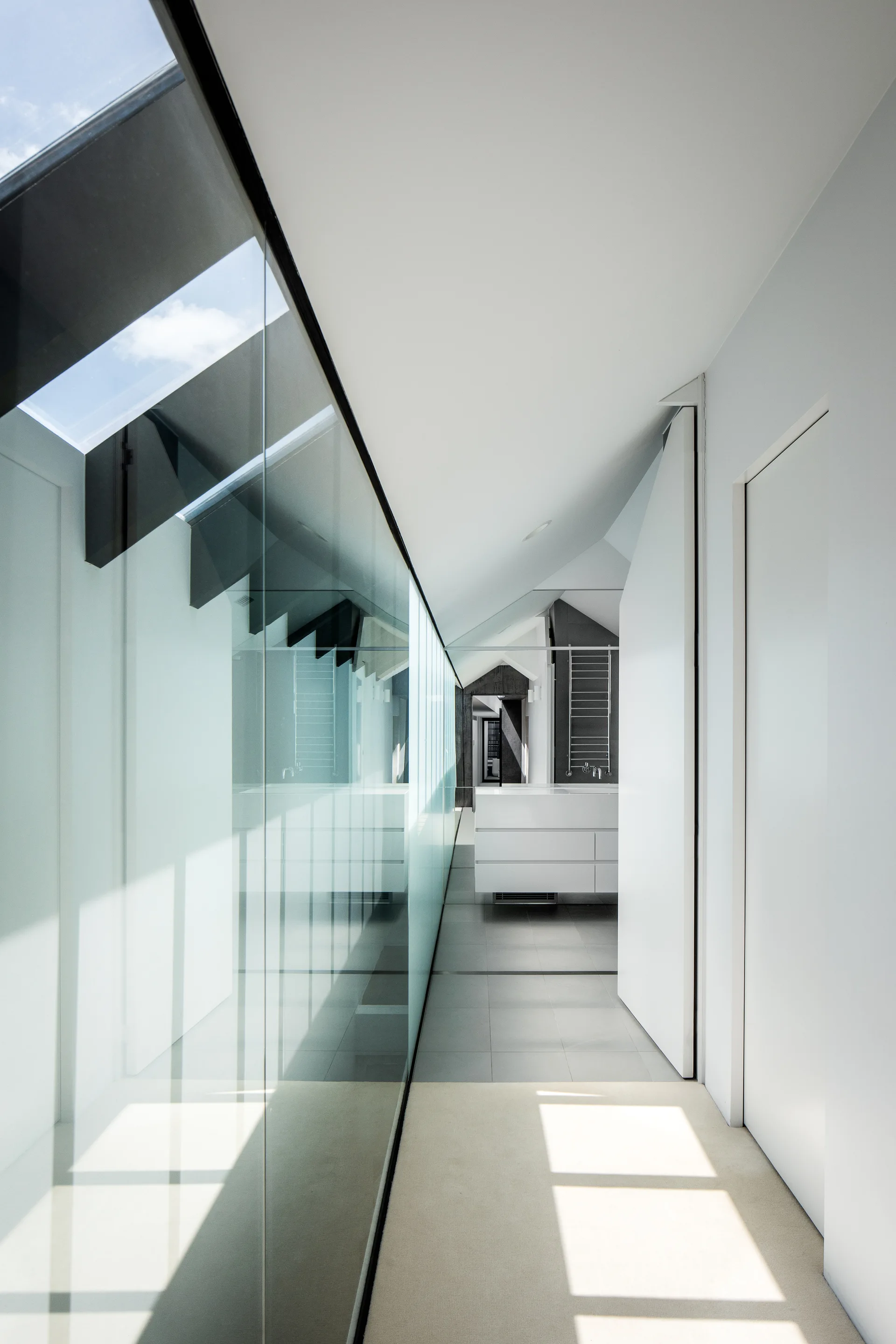 A modern white hallway features a large floor-to-ceiling glass wall on one side, grey floor tiles, and a sloped ceiling with a skylight.