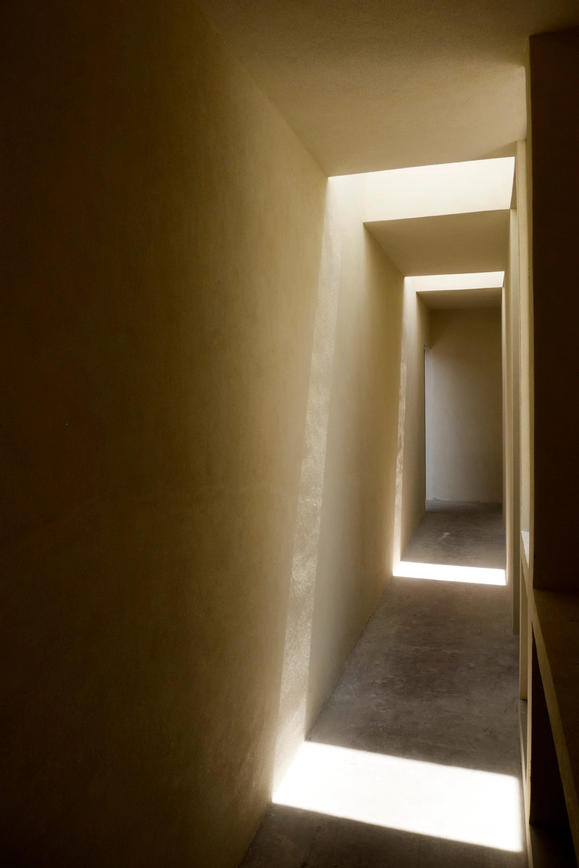 A narrow minimalist corridor with smooth beige plaster walls and concrete floors lit by rectangular skylight openings in the ceiling.
