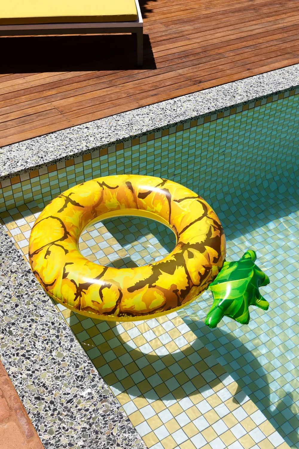 An inflatable pineapple float drifts in a mosaic-tiled swimming pool next to a wooden plank deck and speckled stone coping.