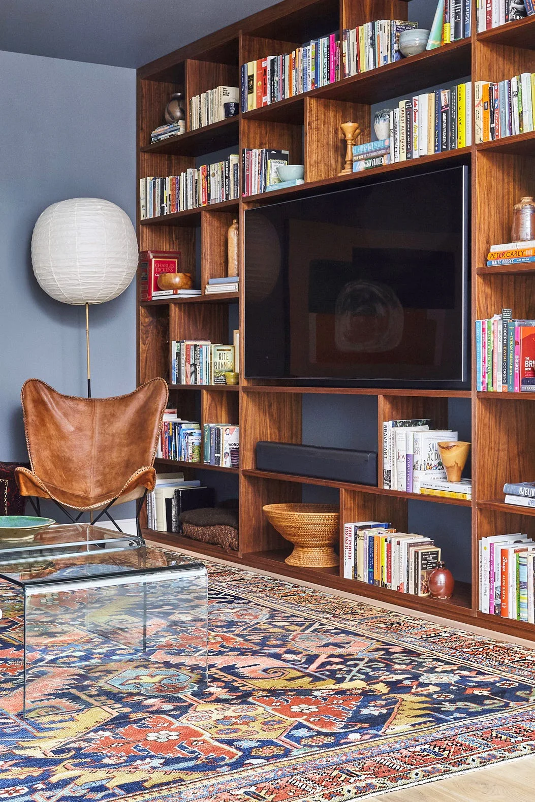 A living room features a large wood built-in bookshelf set against a dark blue wall, a leather butterfly chair, and a colorful patterned area rug.