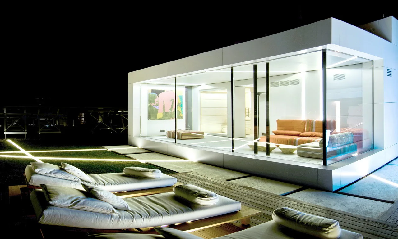 White sun loungers sit on a wooden deck in front of an illuminated modern glass pavilion containing an orange sofa at night.