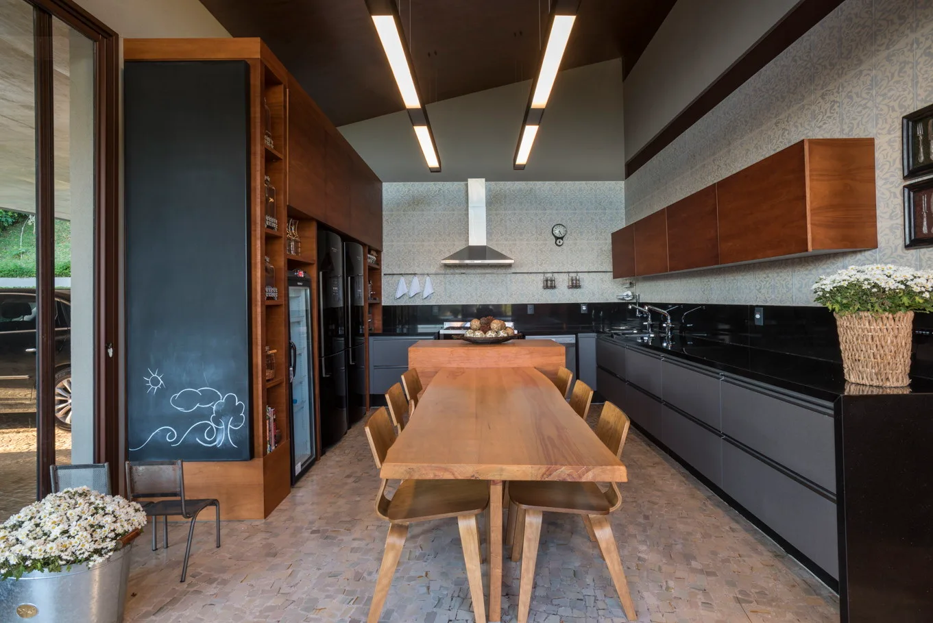 A contemporary kitchen features a long wooden dining table, dark grey lower cabinets, and two linear pendant lights hanging from a dark wood ceiling.