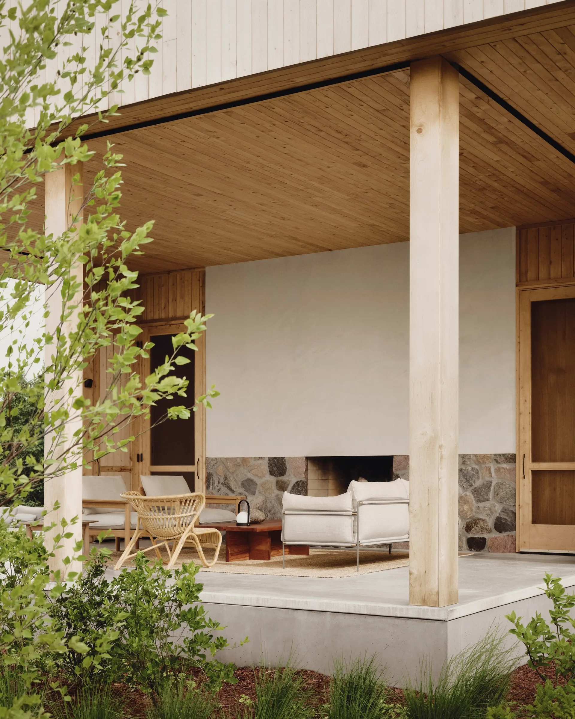 A covered concrete patio features a stone fireplace, wood-plank ceiling, neutral-toned upholstered chairs, and a woven rattan armchair.