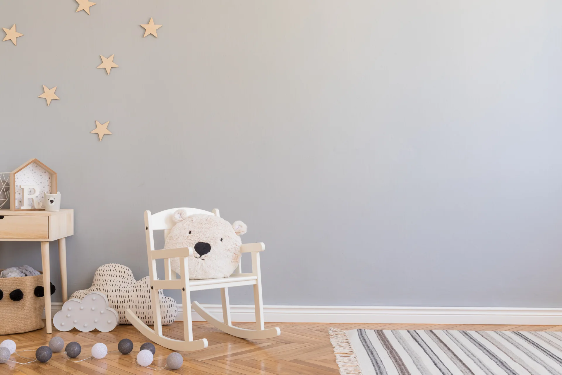 A minimalist nursery corner features a white rocking chair with a bear cushion next to a light wood side table against a pale gray wall.