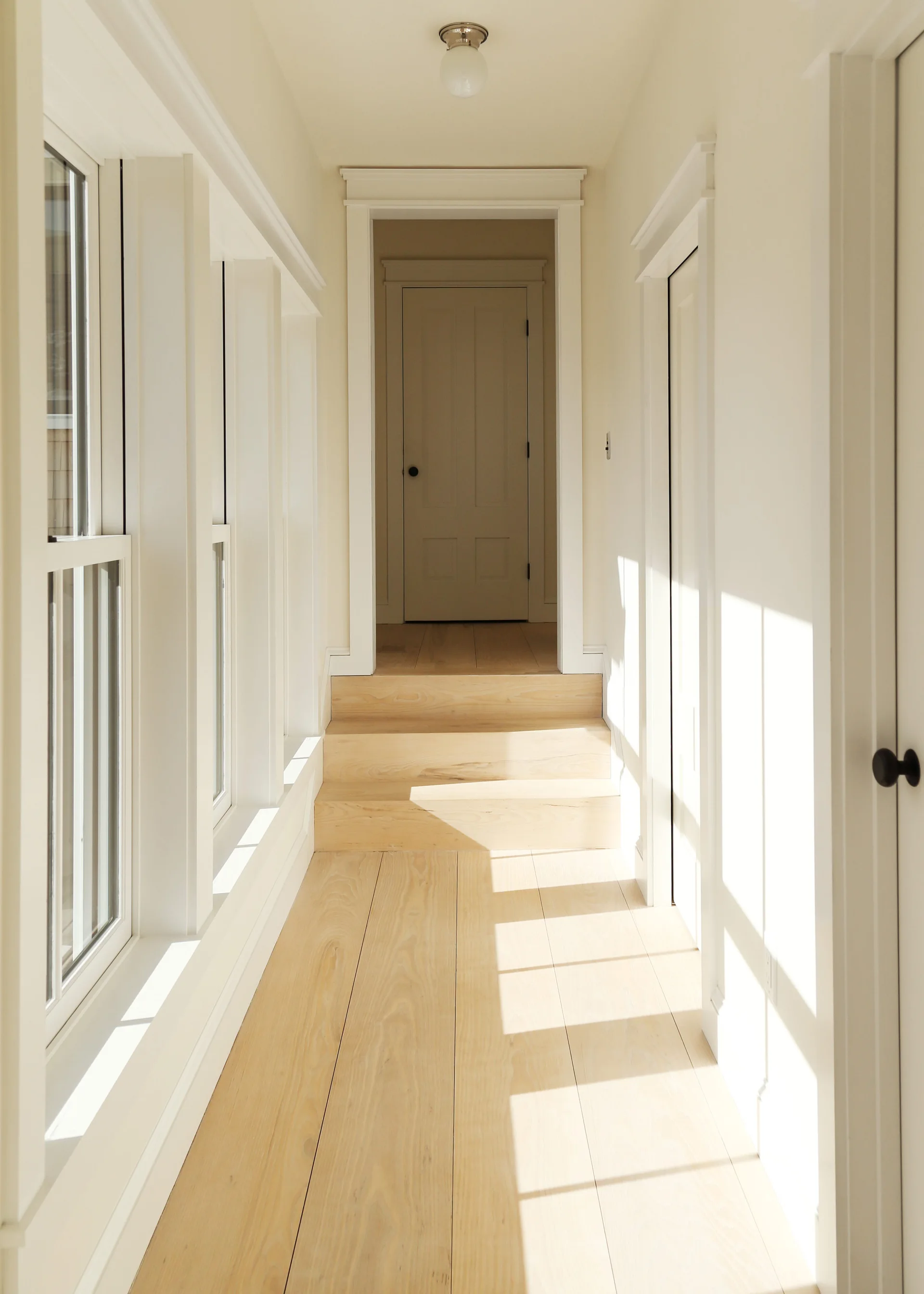 A bright hallway features light wood plank flooring, a small set of wooden steps, white walls with classic trim, and windows casting sunlight shadows.