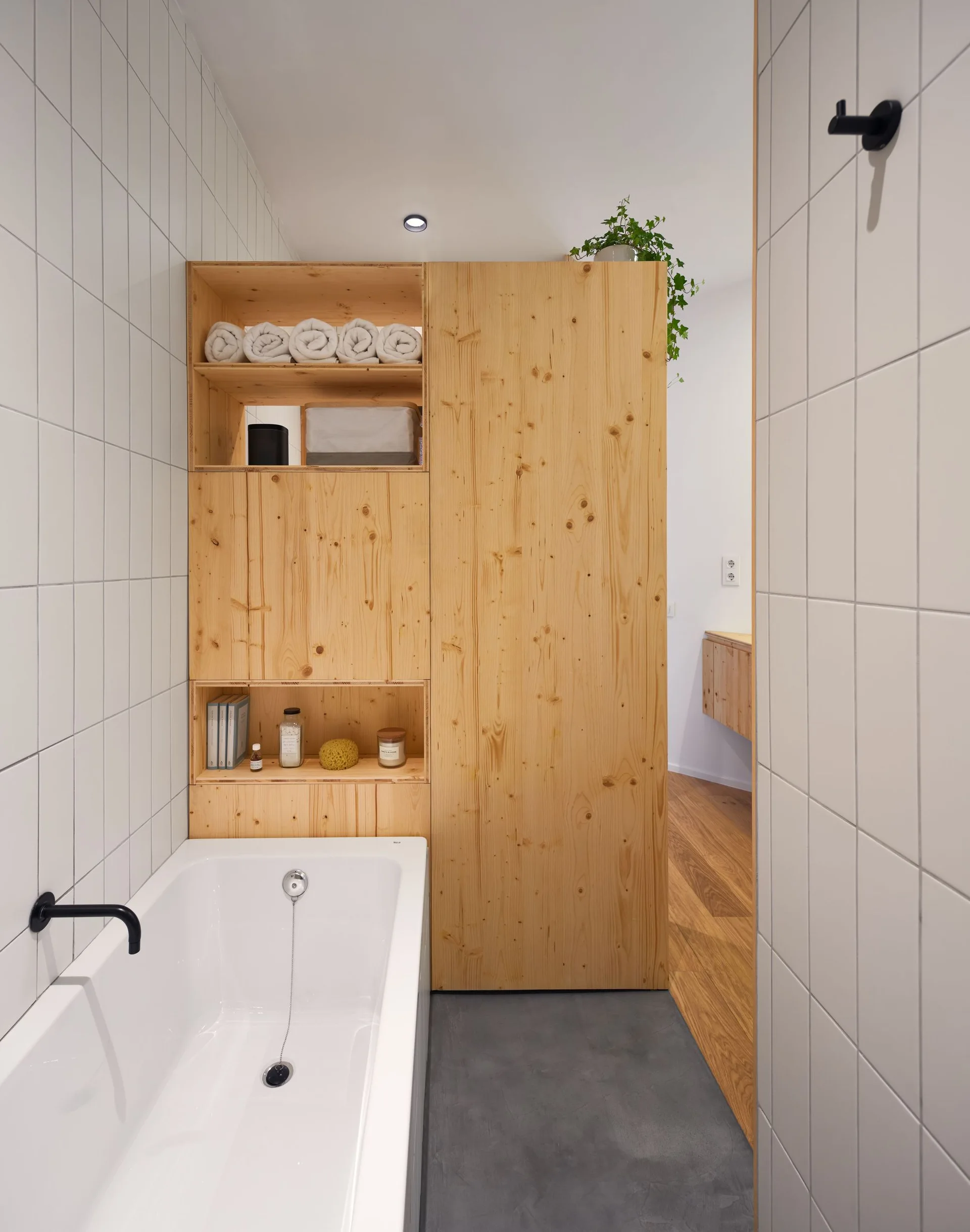 A modern bathroom features a white bathtub, white grid-tiled walls, and a floor-to-ceiling knotty pine cabinet with open shelving.