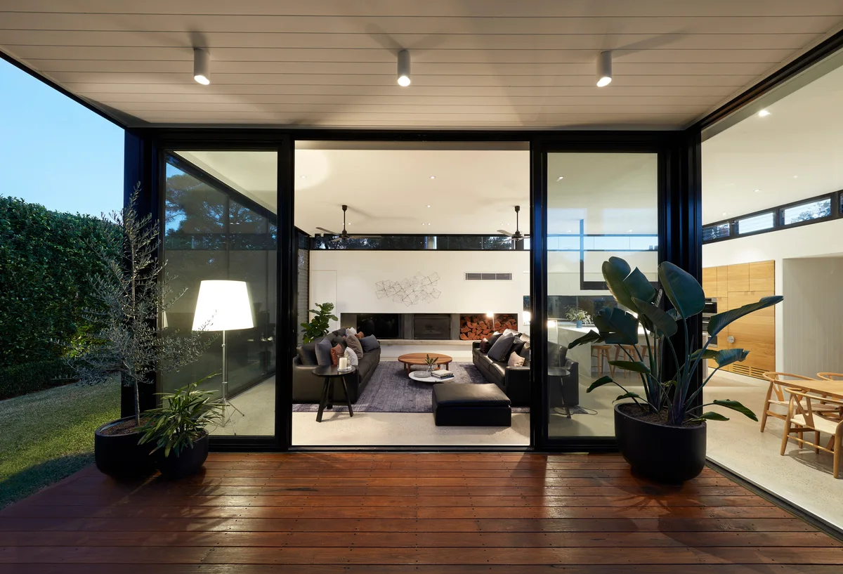 A modern living room with dark leather sofas is seen through large sliding glass doors from an outdoor wooden deck at dusk.