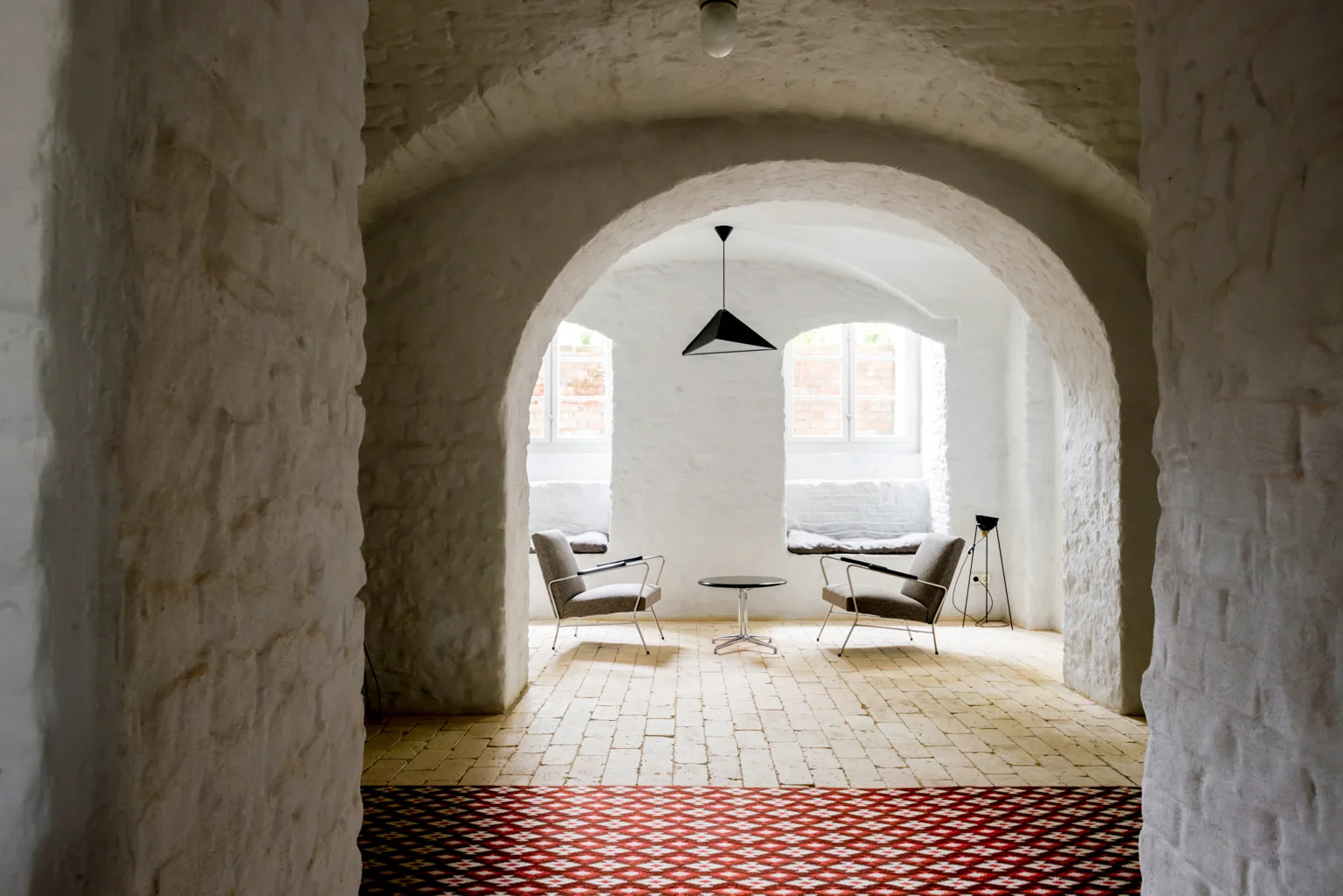Two grey mid-century armchairs and a small black table sit under a whitewashed brick archway with a geometric pendant light and brick floors.