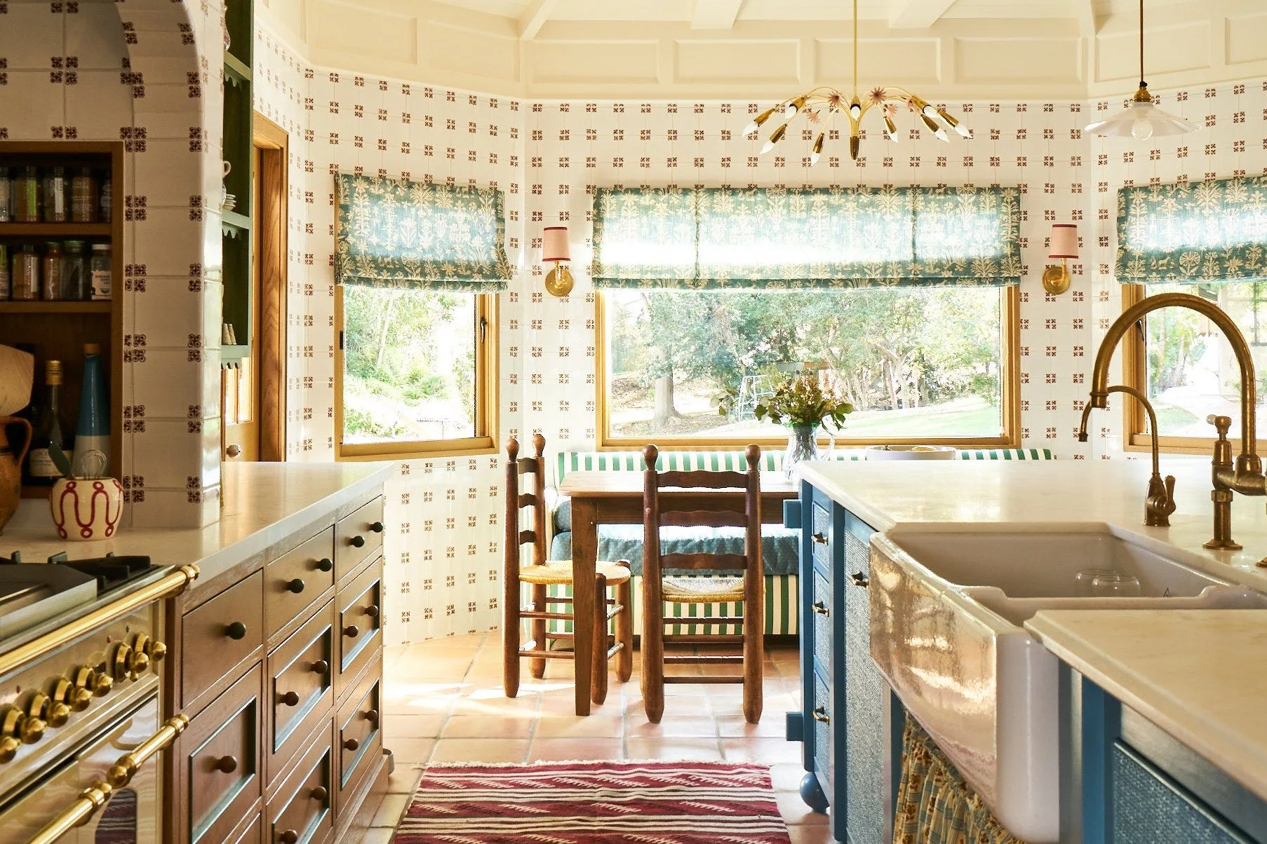 A cottage kitchen features a blue island with a farmhouse sink, patterned wallpaper, and a dining nook with a built-in bench under windows.