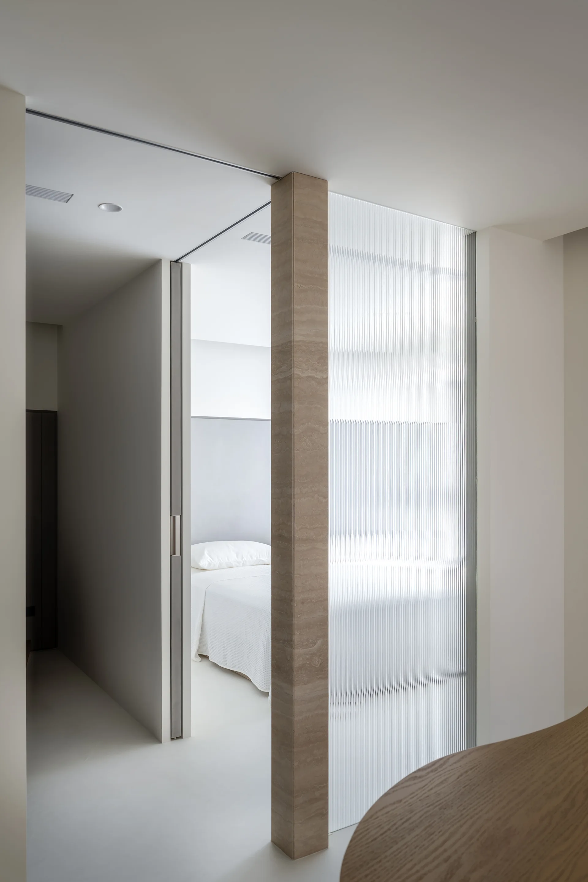A minimalist hallway features a honed stone pillar and a fluted glass partition looking into a bedroom with white bedding.